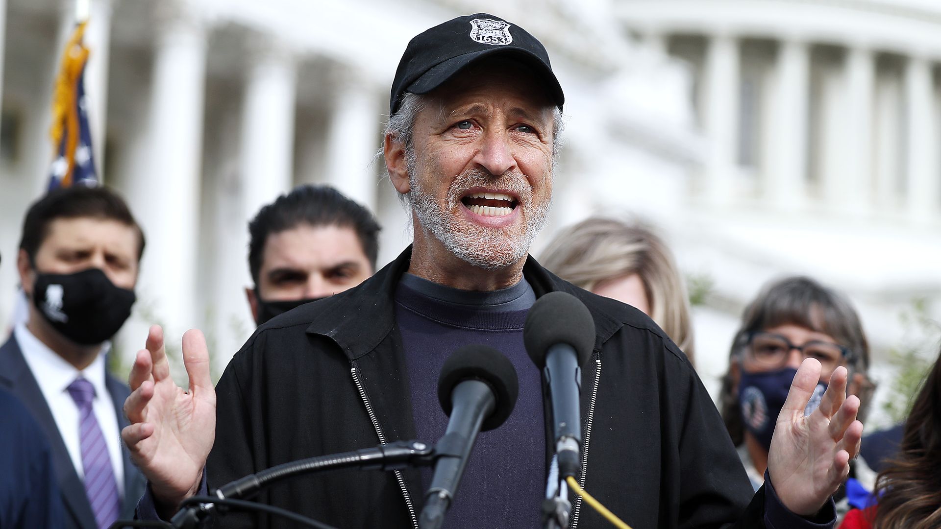 Jon Stewart speaks in front of two microphones while wearing a baseball cap