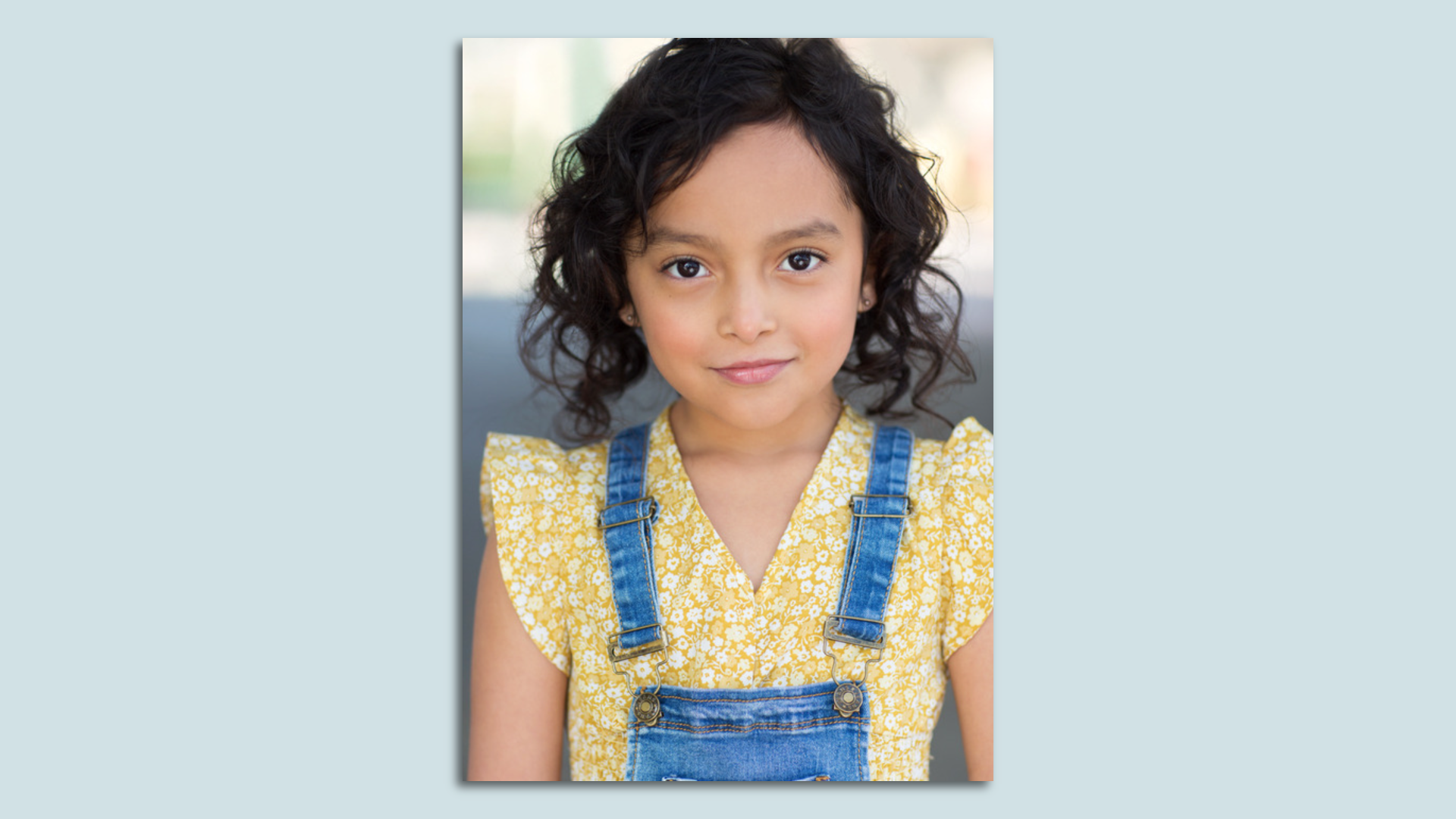 A headshot of a young girl in a yellow shirt and denim overalls.