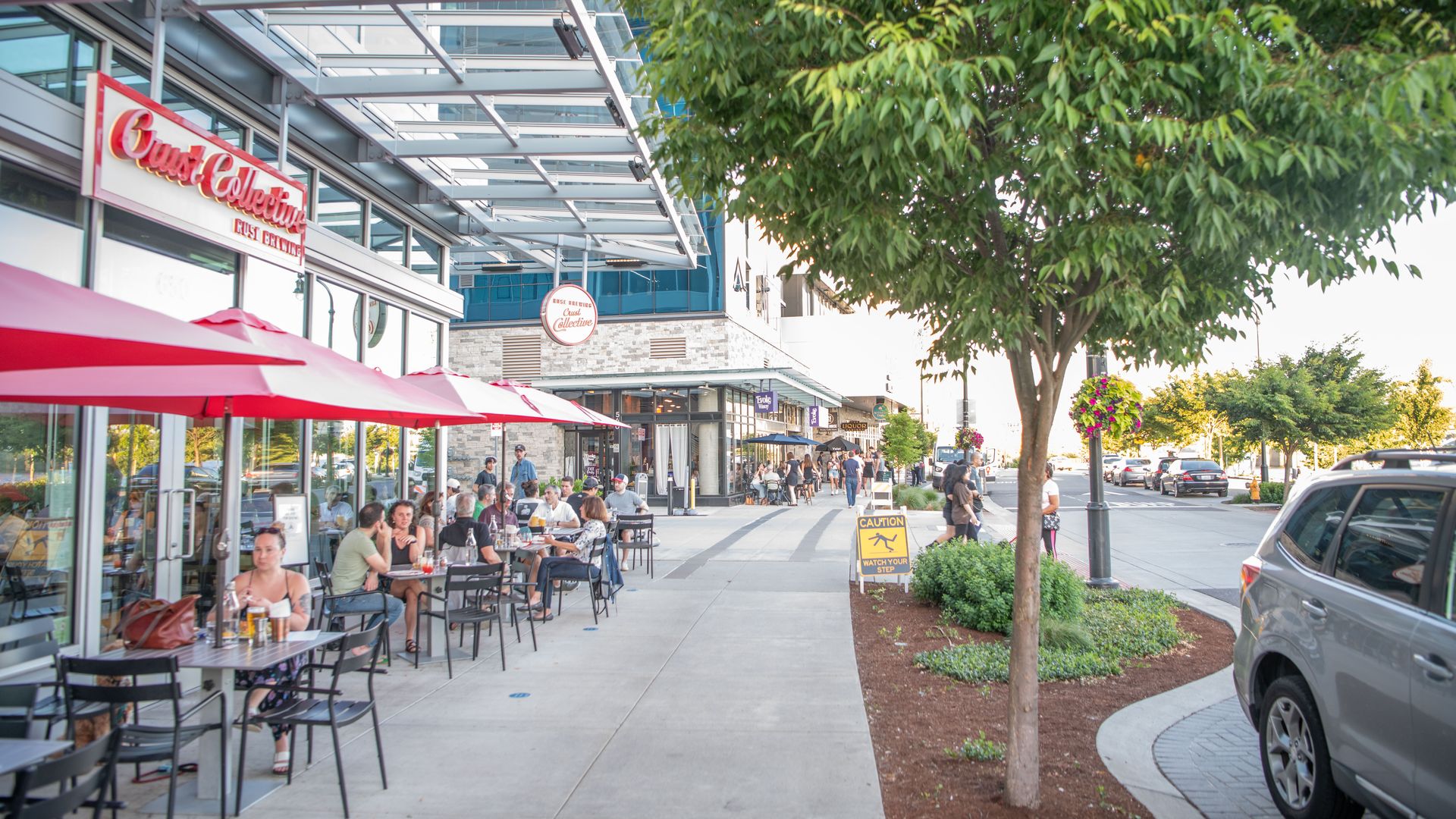Urban sidewalk cafe scene with diners at tables under red-and-white umbrellas, glass storefronts and a metal canopy, a large leafy tree on the right, pedestrians walking, and cars parked nearby.