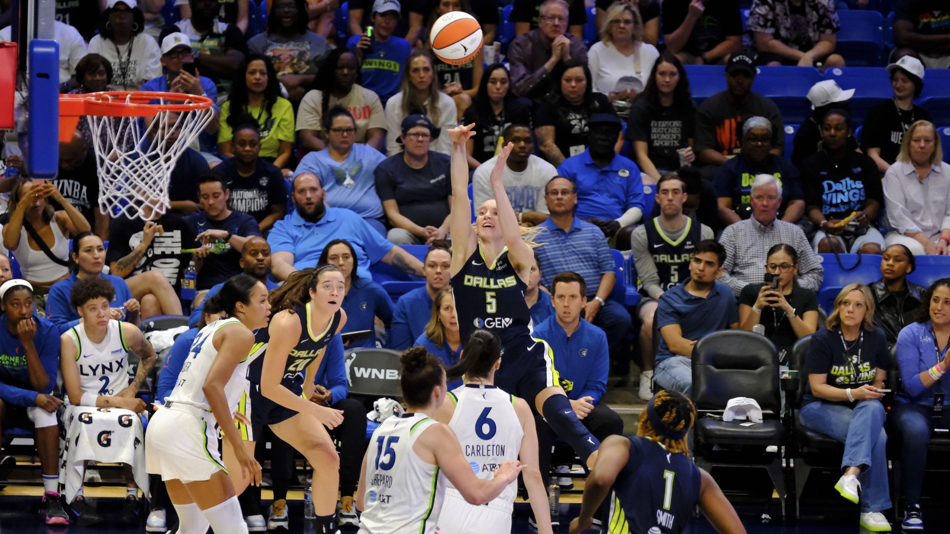 A woman wearing black shoots a ball into a basket with players in white trying to block her