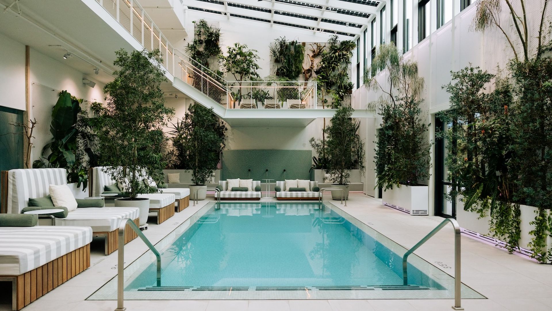 Indoor swimming pool with light blue water, surrounded by white and wood lounge chairs with green cushions, tall leafy green plants, and a white glass ceiling with a ceiling fan.