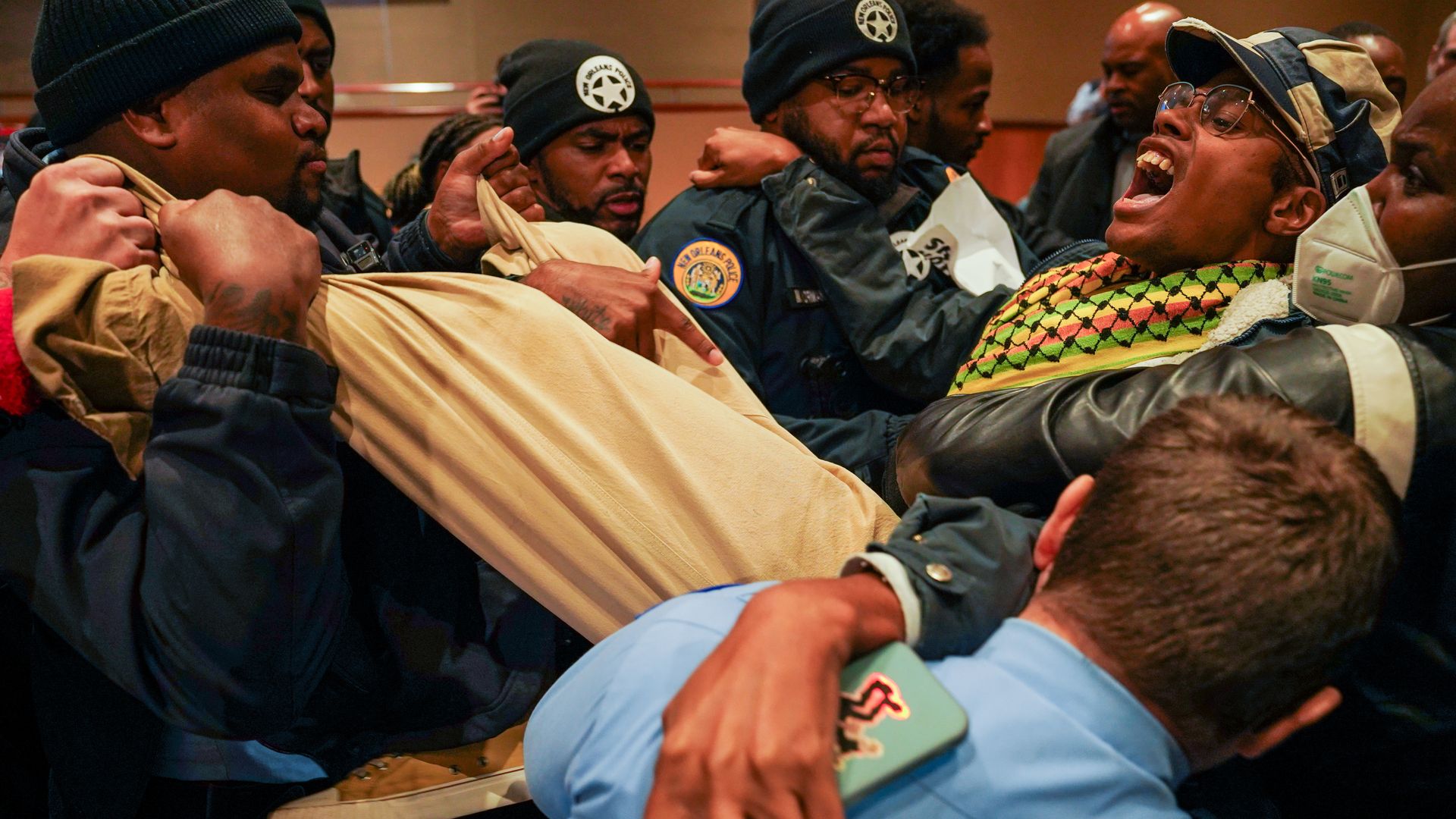 A person wearing a hat and multicolored scarf yells while they are carried by at least five New Orleans Police officers.