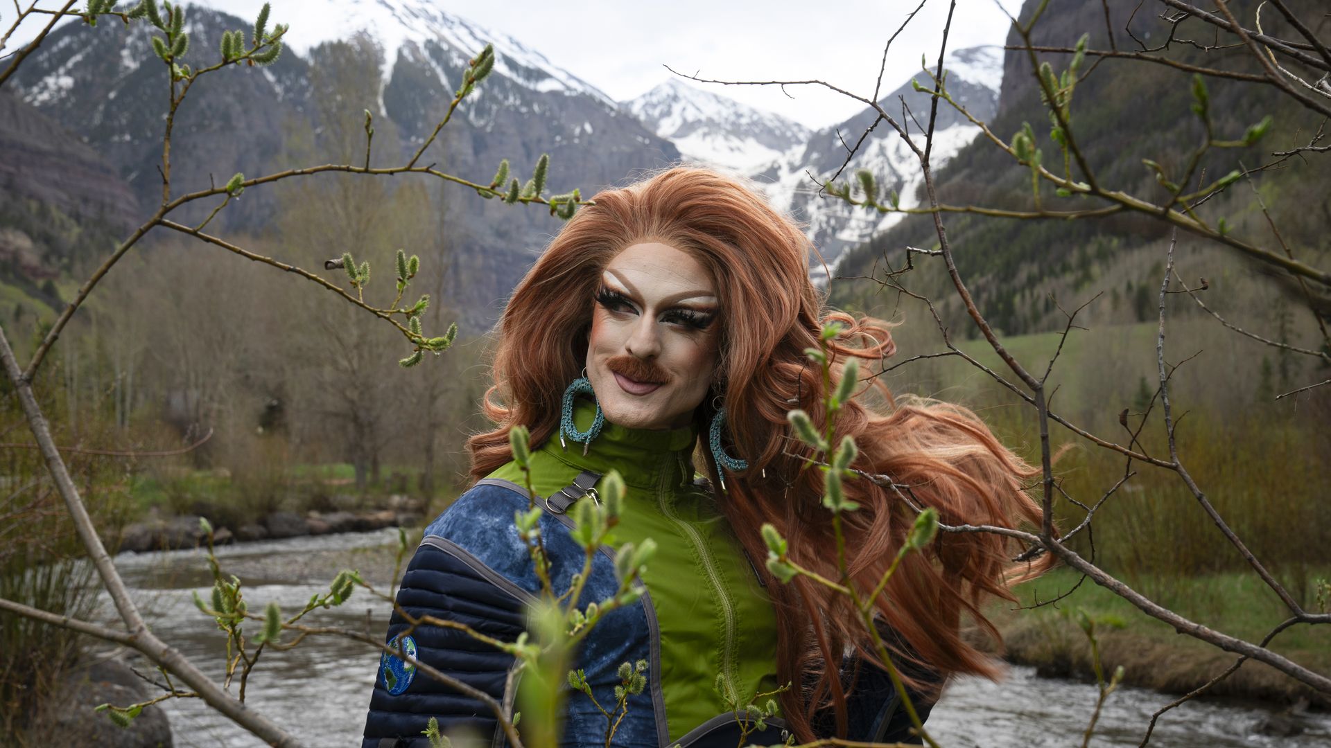 Person with long red hair, bold makeup, wearing a green vest and turquoise earrings, stands among branches with a snowy mountain valley and river in the background.