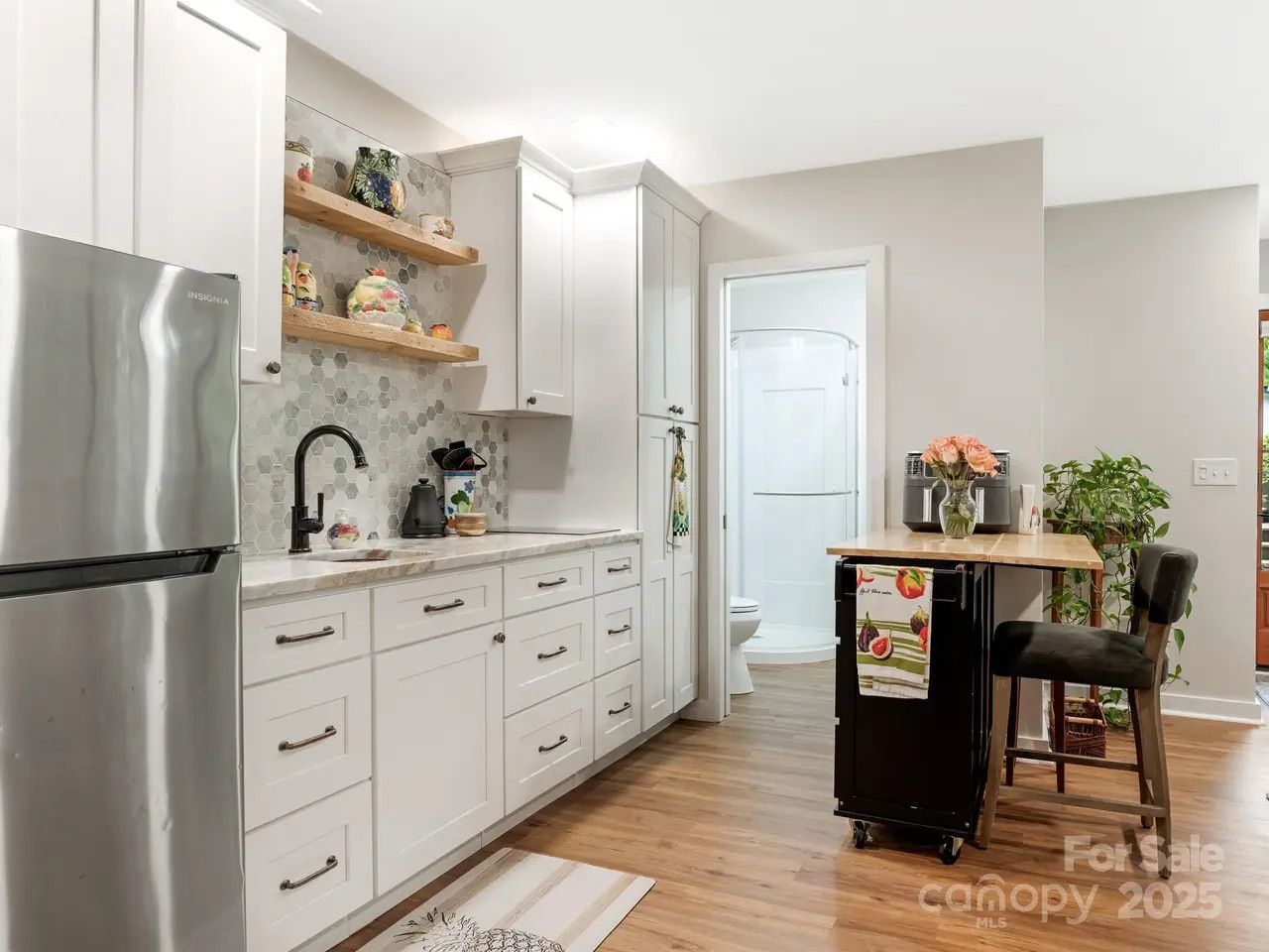 Bright kitchen with white cabinets, hexagonal tile backsplash, stainless fridge, wooden floor, small black island with flowers, chair, and open door to bathroom with glass shower.
