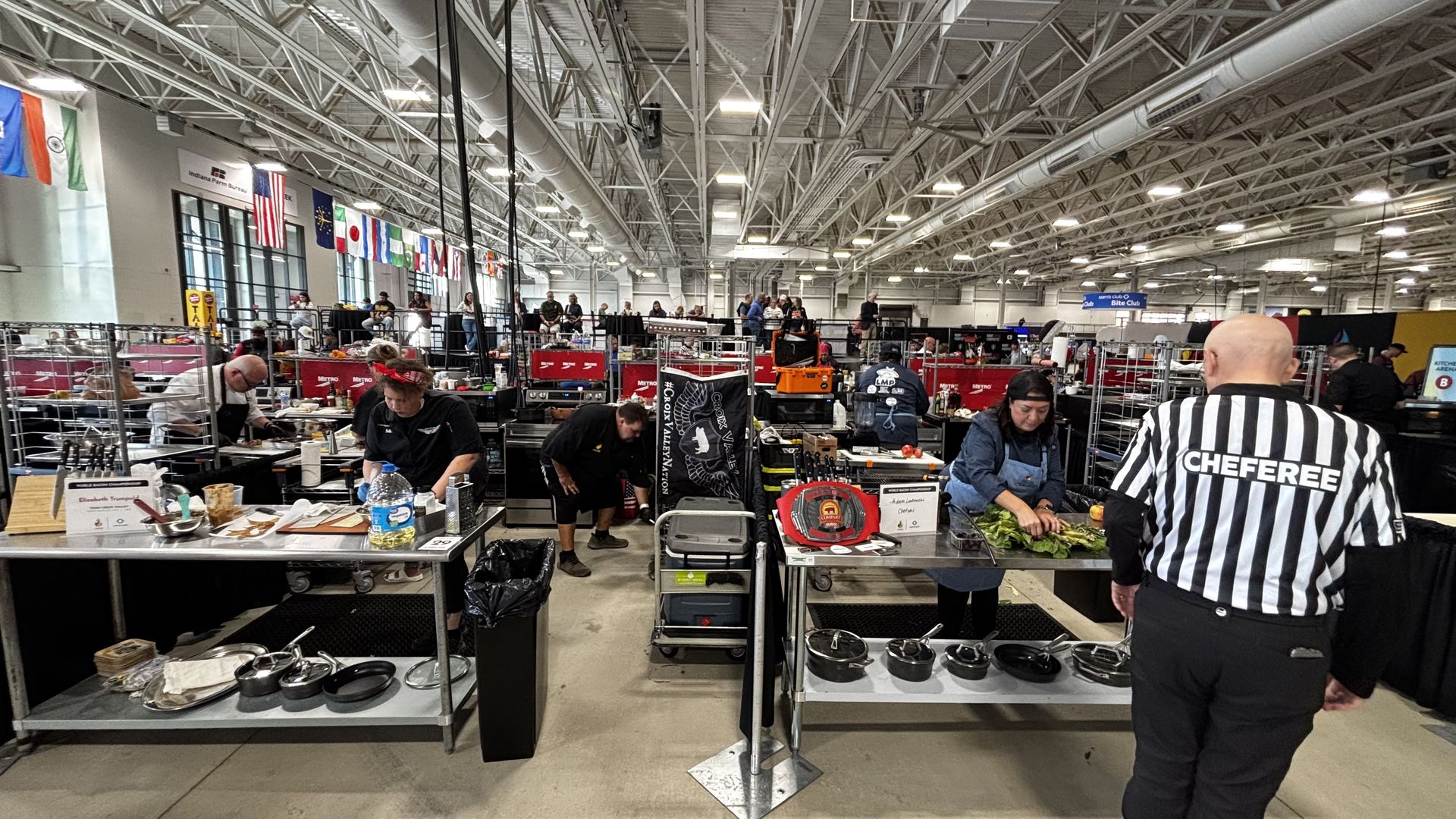 Indoor cooking competition with chefs working at stainless steel tables filled with ingredients and utensils. A referee in a black-and-white striped shirt walks nearby. Flags hang above.
