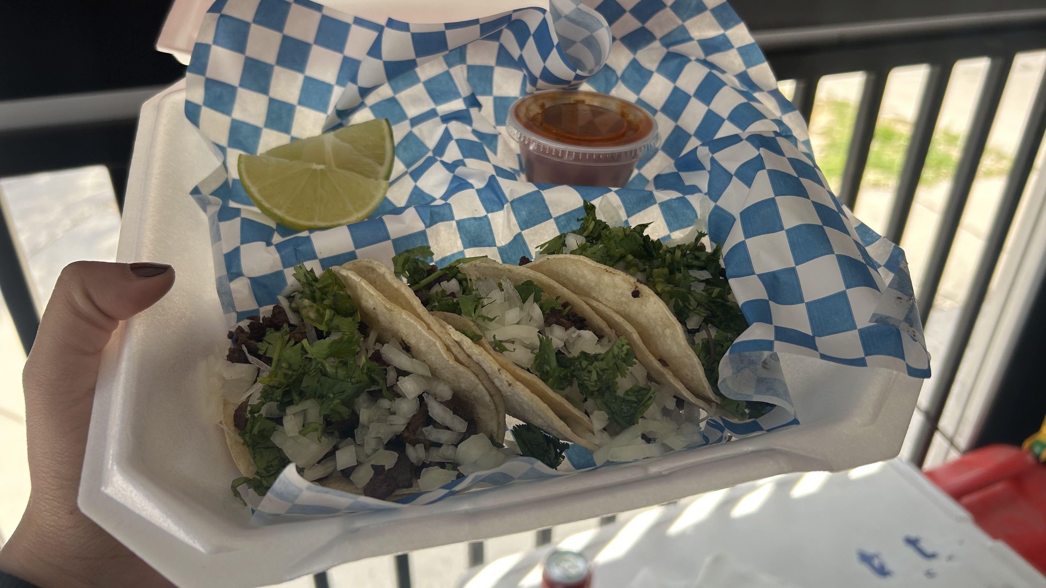 Two tacos with chopped onions and cilantro in a blue-and-white checkered liner, lime wedge, and a small cup of red salsa; a hand holds the takeout tray outdoors.