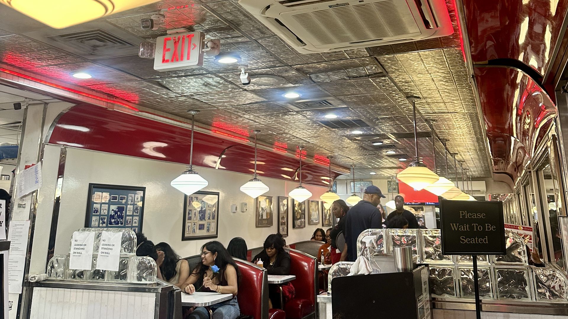 Interior of a busy diner with red booths, silver patterned ceiling, hanging lamps, red neon lighting, an air conditioner, and a sign saying "Please Wait To Be Seated."