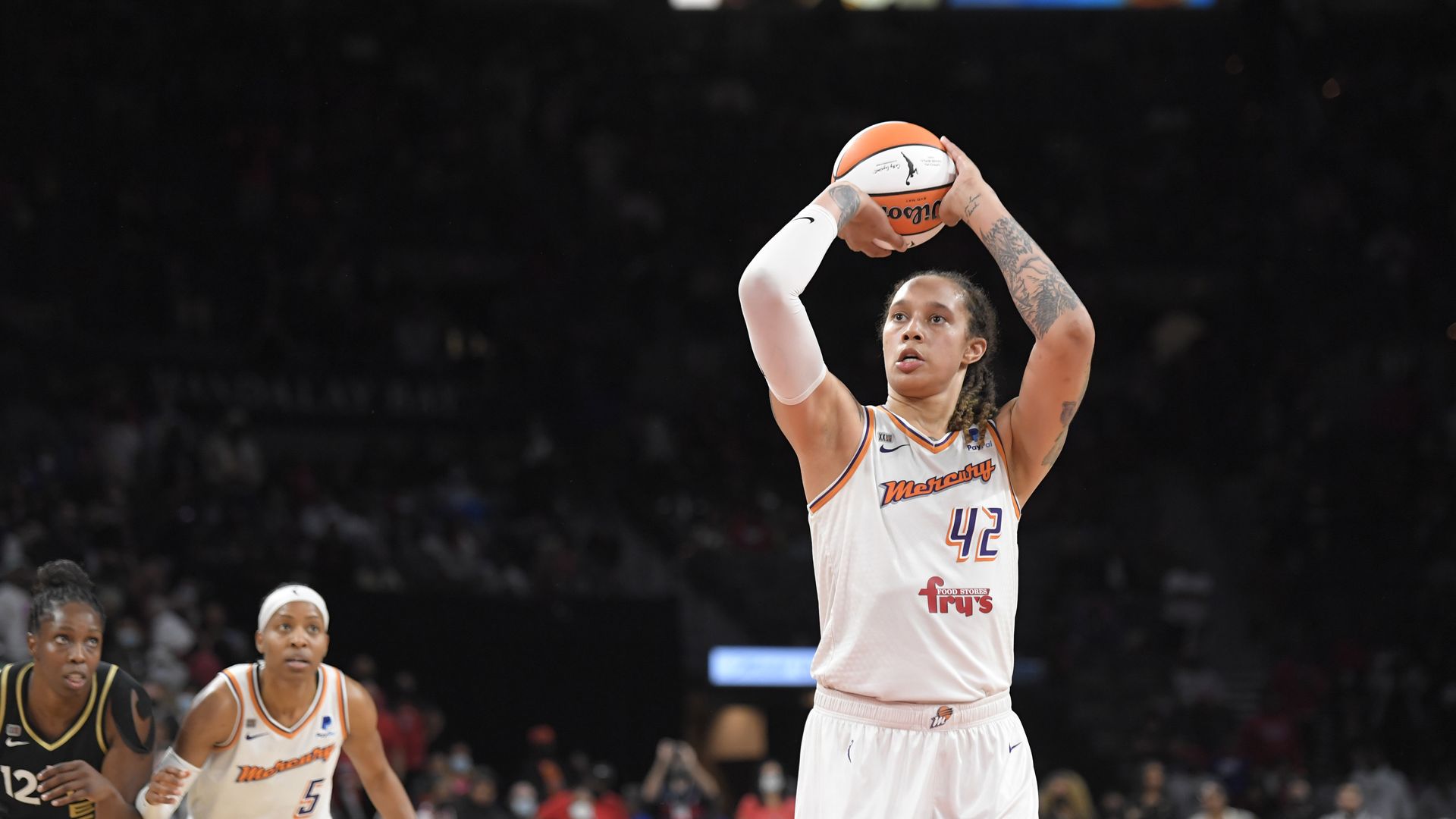 Photo of Brittney Griner holding aiming a basketball in the air on a court as she wears a white and orange Phoenix Mercury uniform