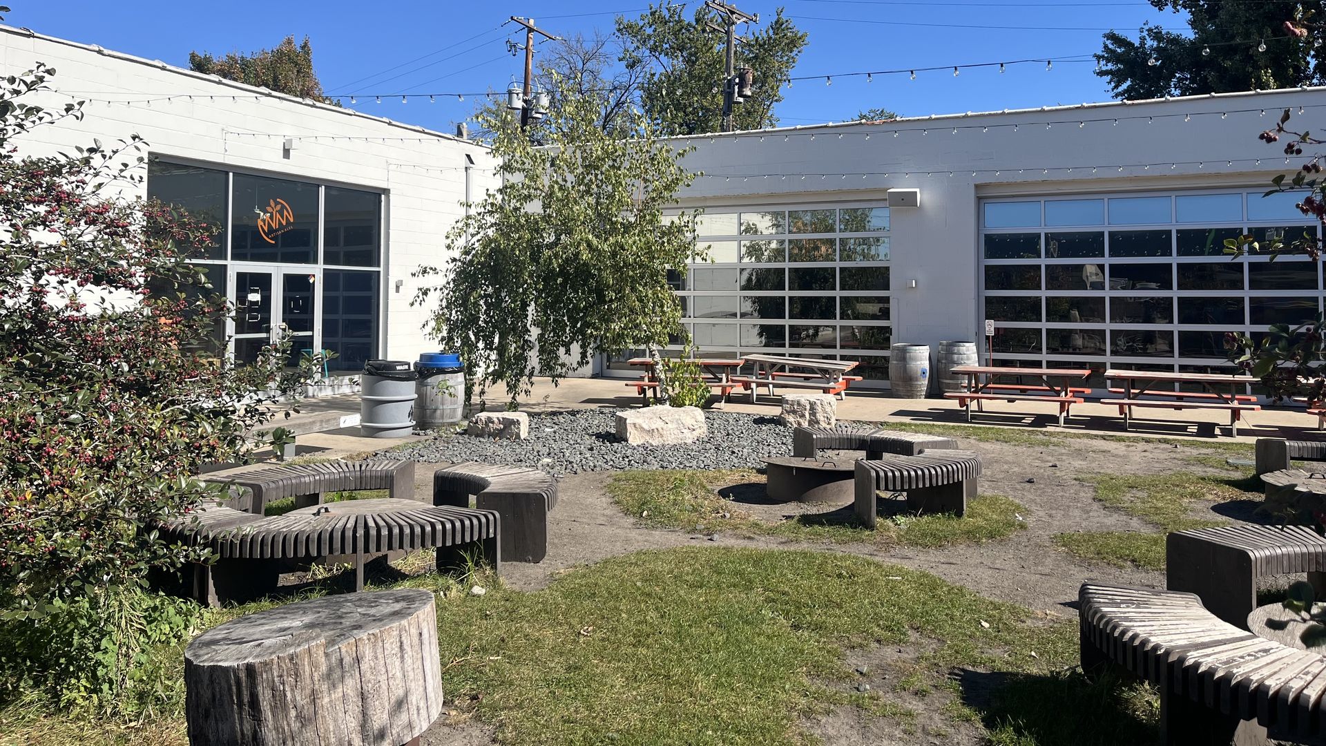 Outdoor seating area with curved wooden benches around a fire pit, picnic tables, string lights overhead, white building with large glass doors, and a clear blue sky.