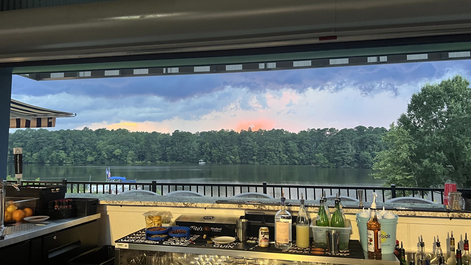 Bar counter in the foreground with bottles, lemons, and mixers; overlooks a lake with a tree-lined shore beneath a cloudy sky and a pink sunset glow.