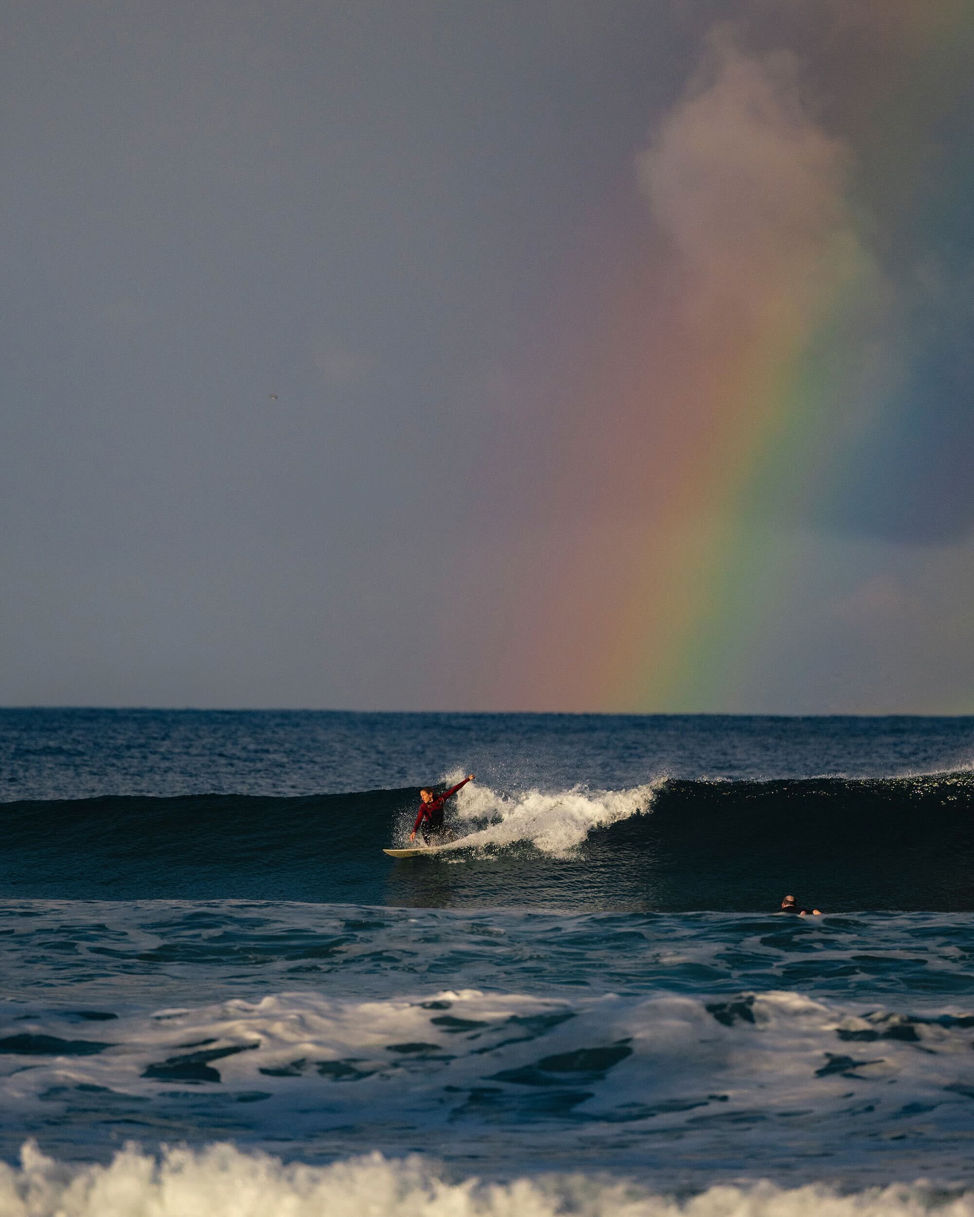 Surfer in a red wetsuit rides a wave on a dark blue ocean with a partial rainbow visible against a cloudy sky in the background.