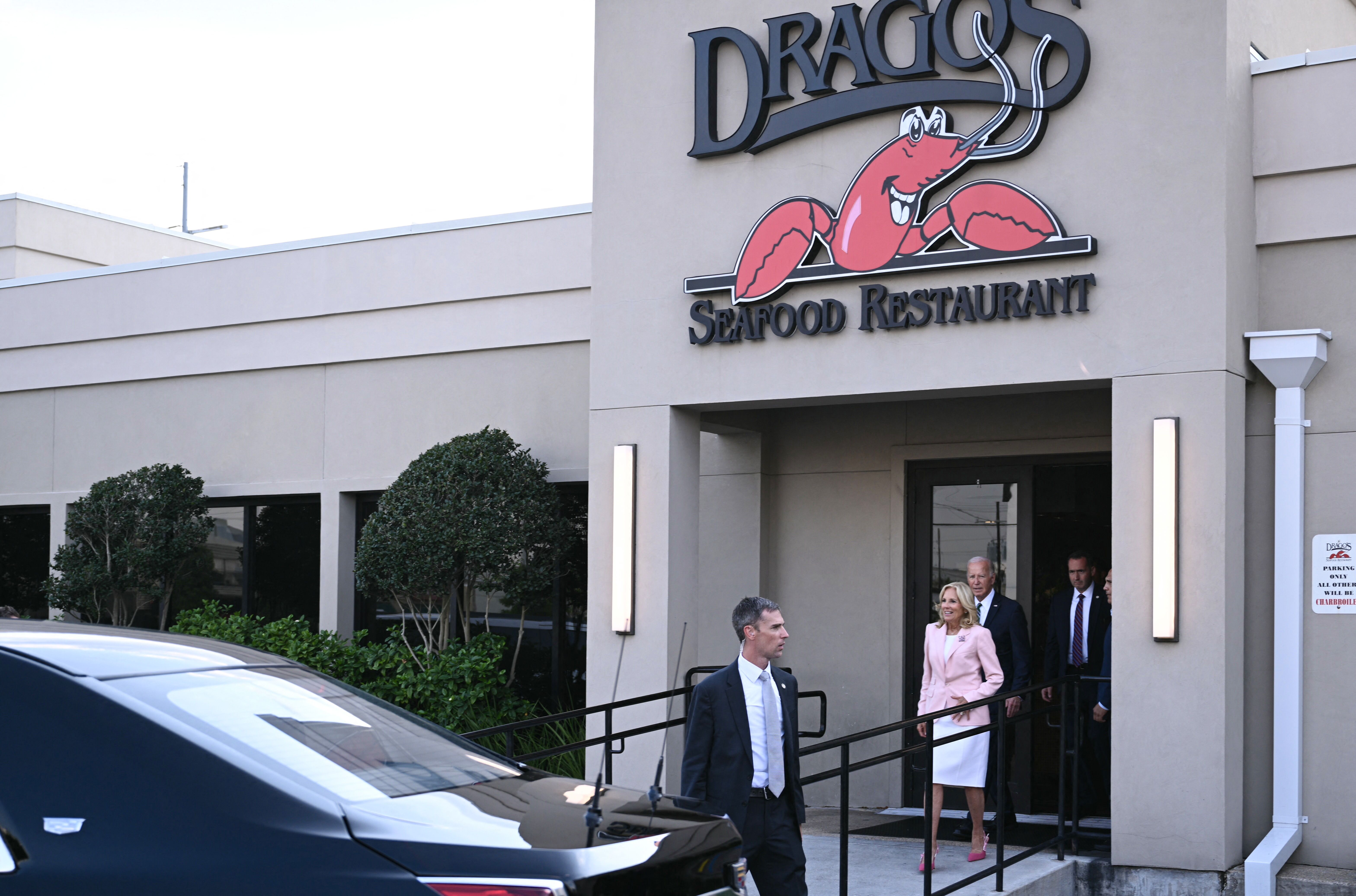 Jill and Joe Biden walk out of Drago's Seafood Restaurant. Another man in a suit walks in front of them. The restaurant's name is on a sign above the door with a cartoon crawfish image.