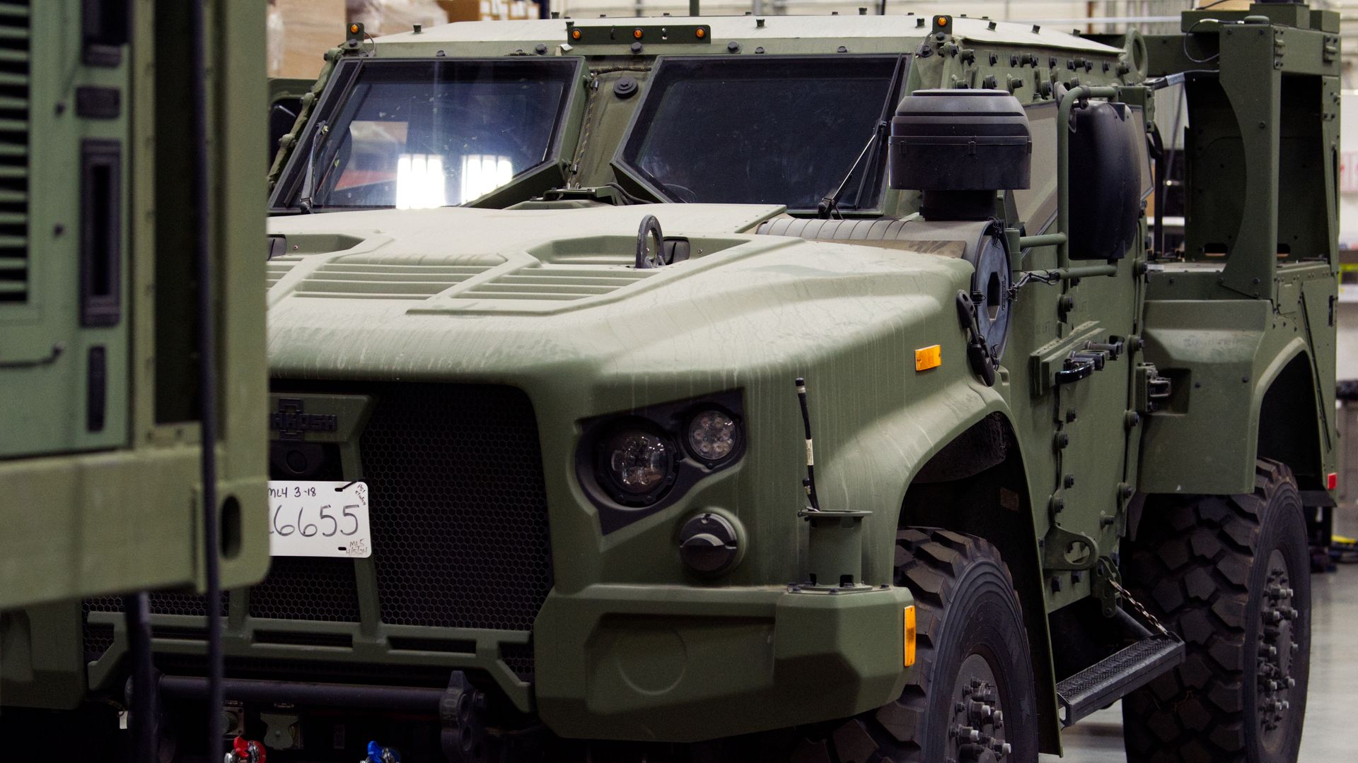 Olive-green military armored vehicle with angled front windows and large rugged tires, parked in a warehouse. Cardboard boxes are seen in the foreground.