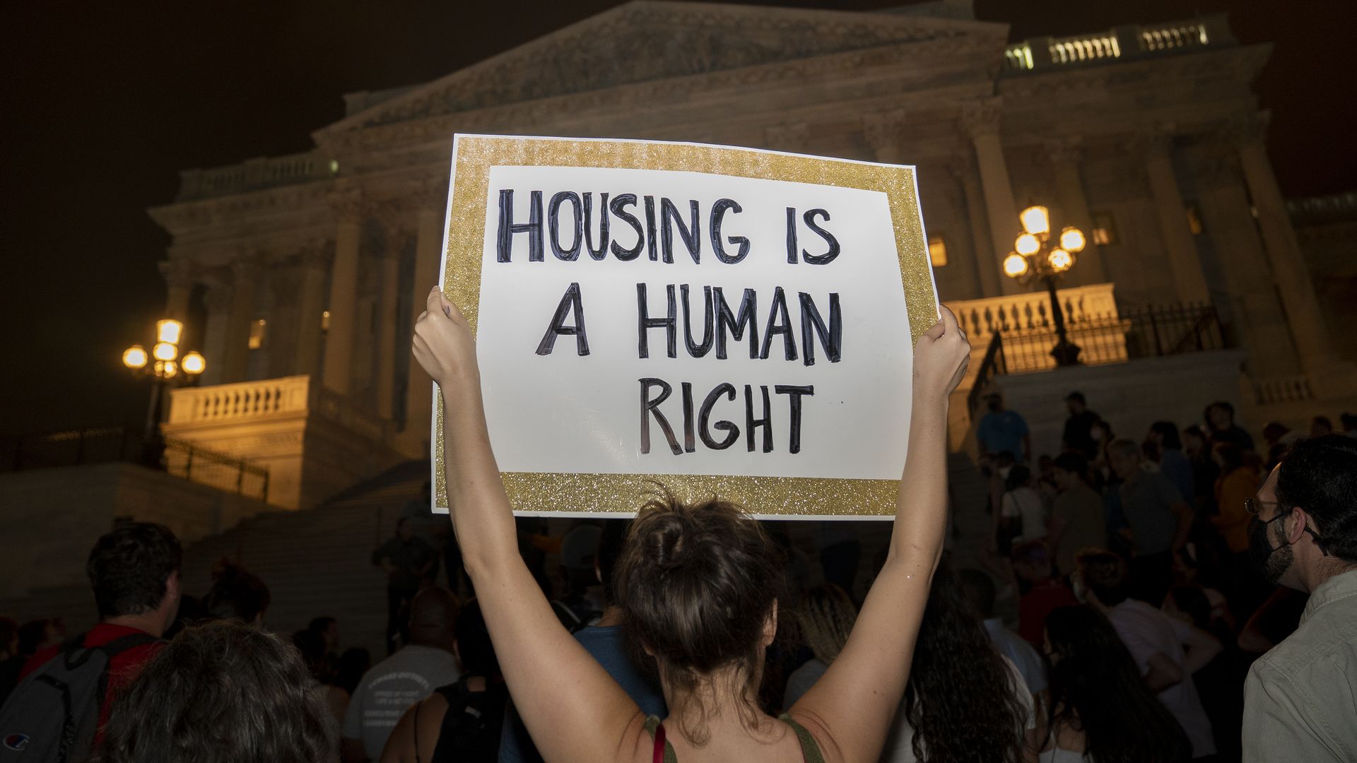Photo of a person holding up a sign that says "Housing is a human right" in front of a building int he nighttime