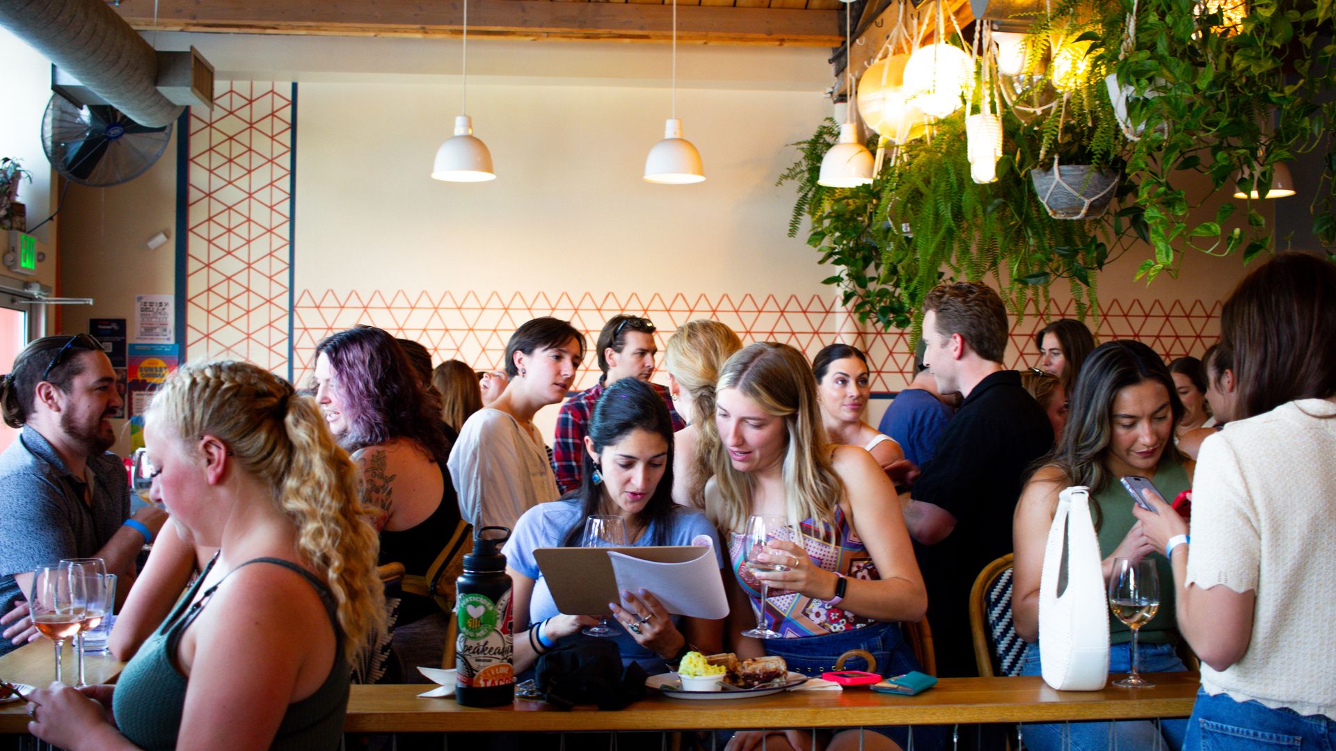 Crowded cafe scene: long wooden counter, groups chatting, two women study a menu at the bar, wine glasses and plates nearby, hanging green plants, walls with orange patterns and pendant lights.