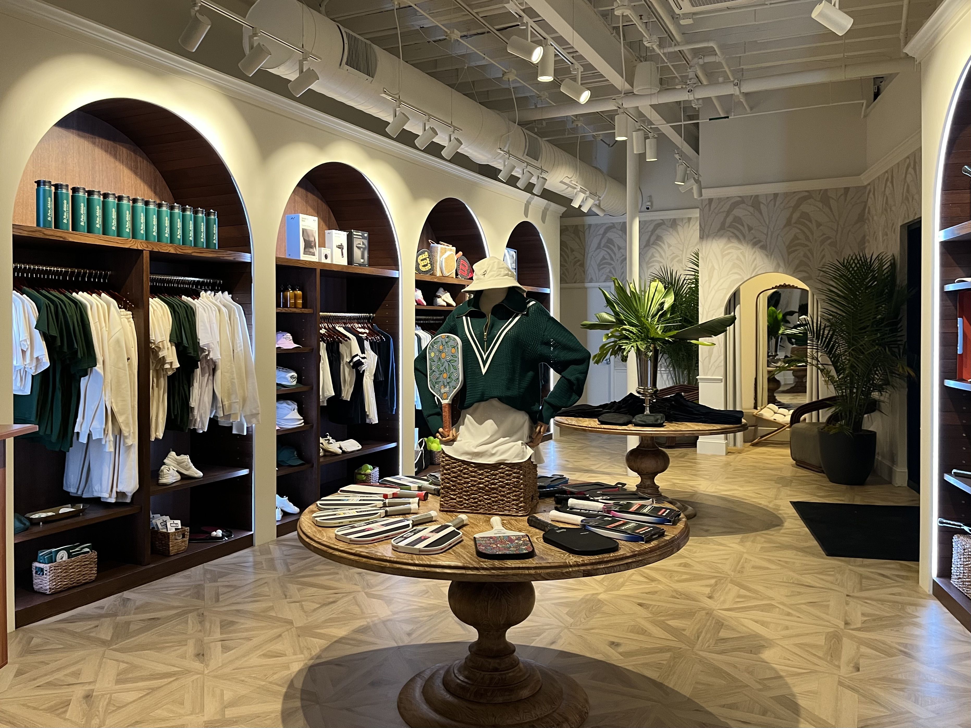 Bright sports apparel store interior with wooden round tables showcasing paddles, mannequin dressed in green sweater and hat, arched shelves holding clothes and accessories, plants and large mirror.