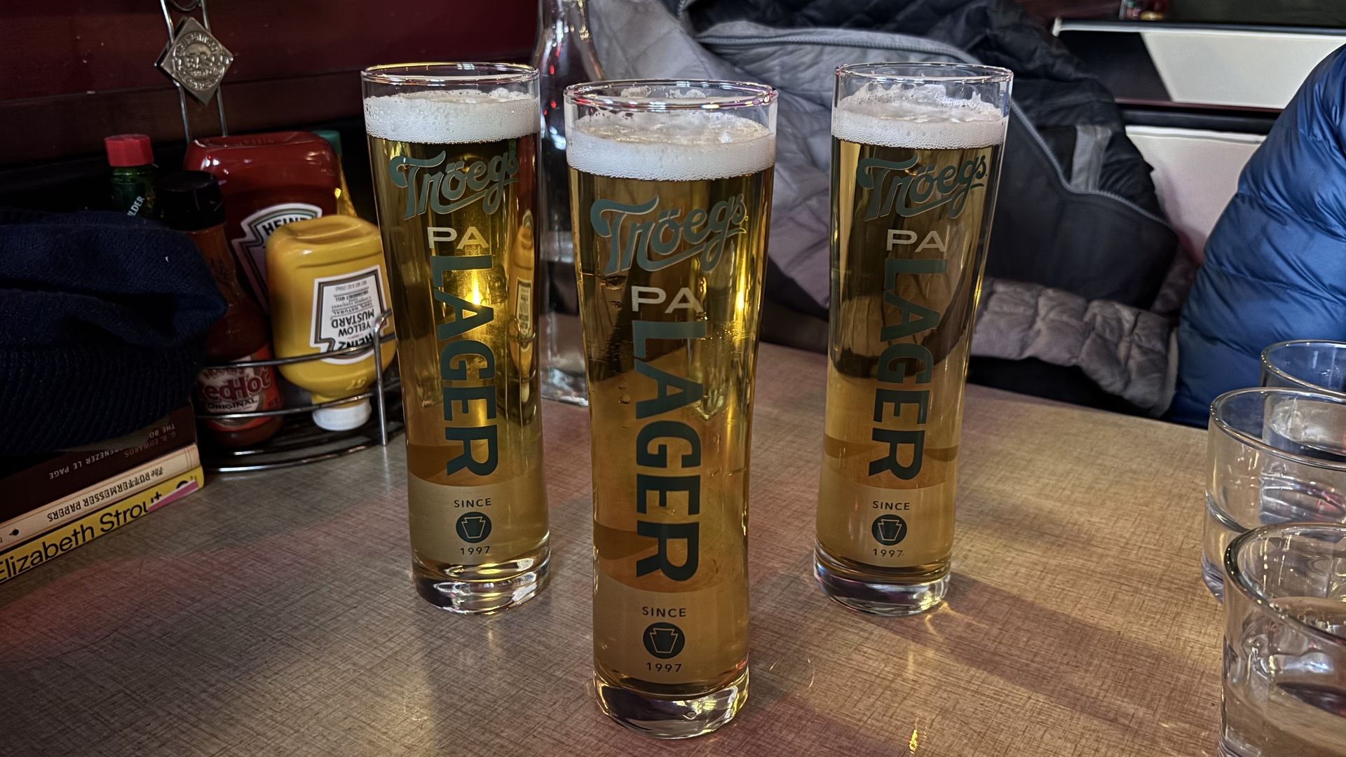 Three tall Troegs beer glasses on a wooden table, pale golden liquid with foamy heads, glassware labeled PA and Since 1997. Background shows condiments, jackets, and a gloved arm on the right.