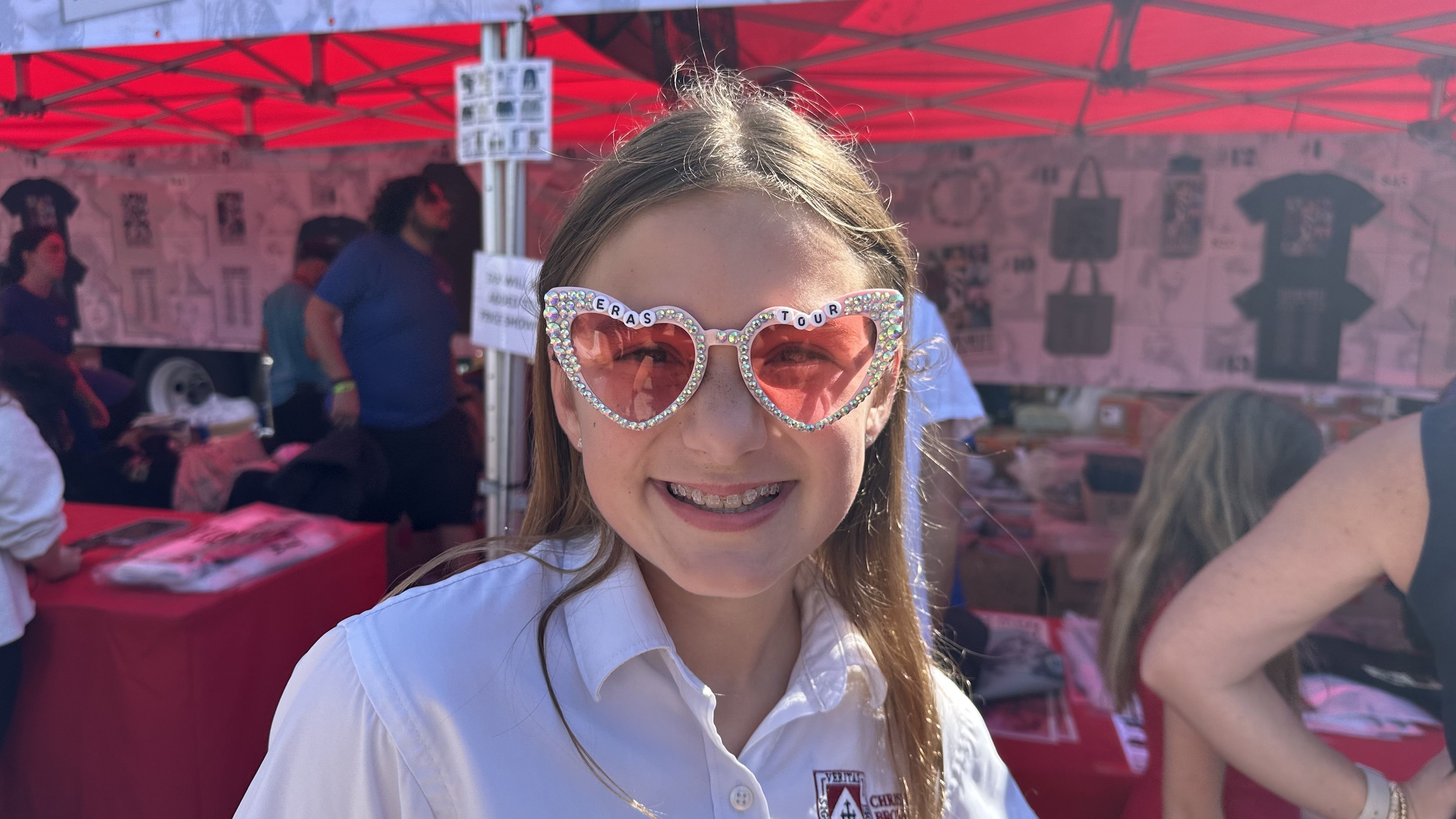 A young girl smiles while wearing pink heart-shaped sunglasses that say ERAS TOUR across the top.