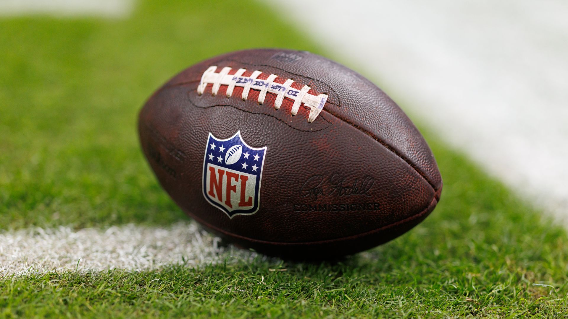 A close-up of a brown leather NFL football with white laces and the NFL shield logo, resting on green grass with a blurred field in the background.