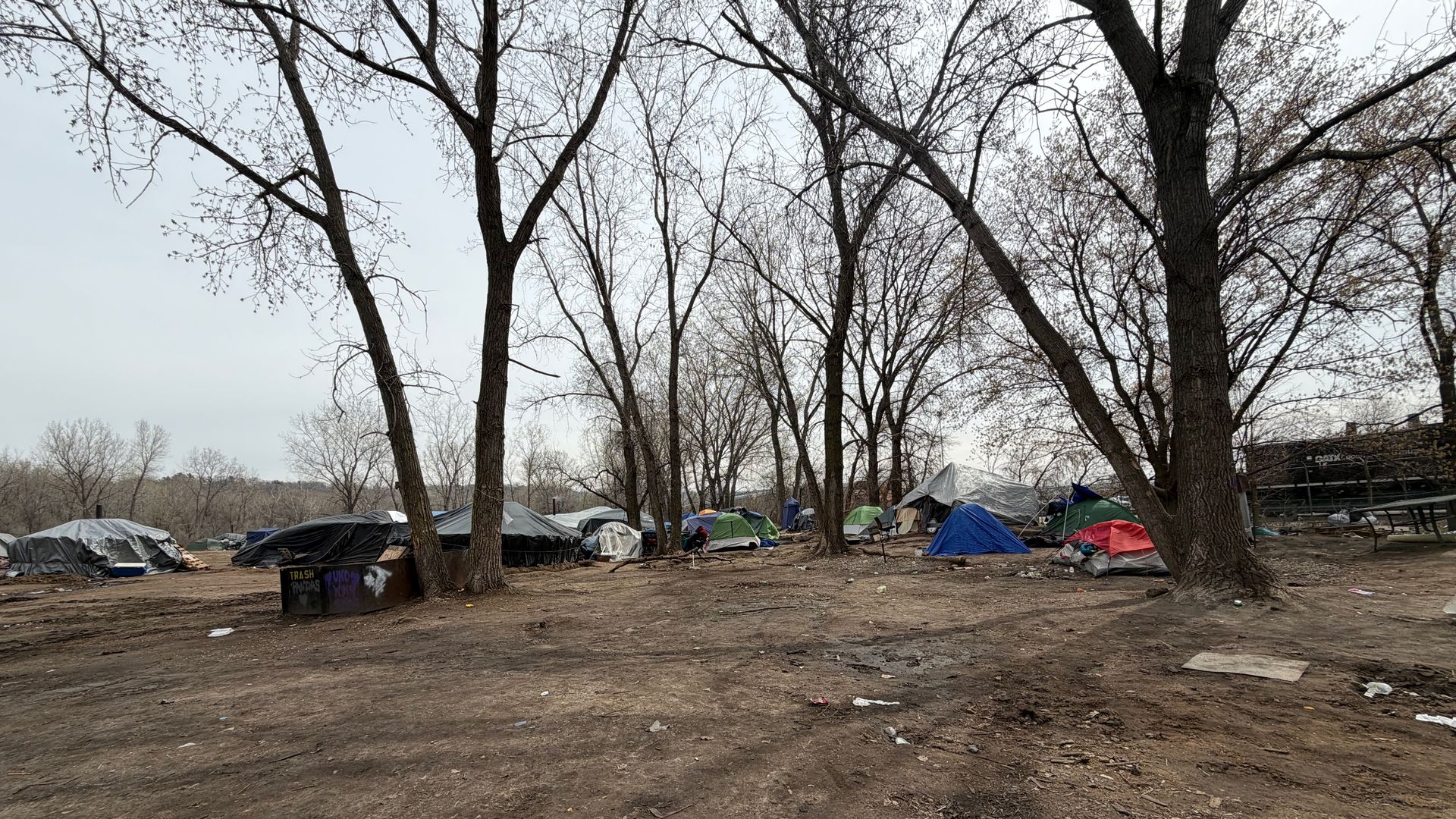 A row of leafless trees over a dirt clearing, with colorful tents pitched beneath. Trash and litter scattered on the ground, a bleak overcast sky above.