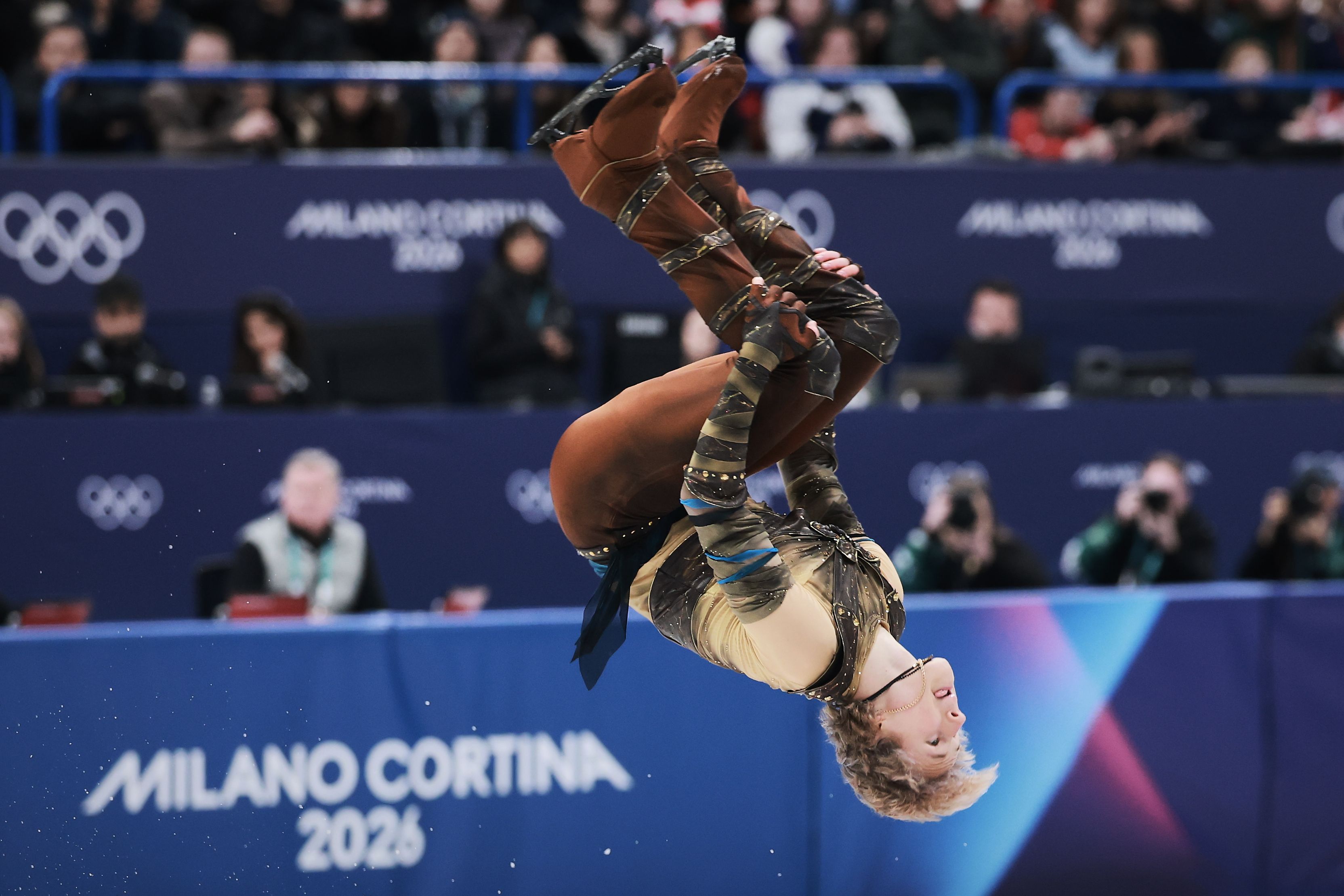 Figure skater Ilia Malinin in brown and gold costume performing a mid-air flip during a competition with spectators and photographers in the background at Milano Cortina 2026 Olympic Games.