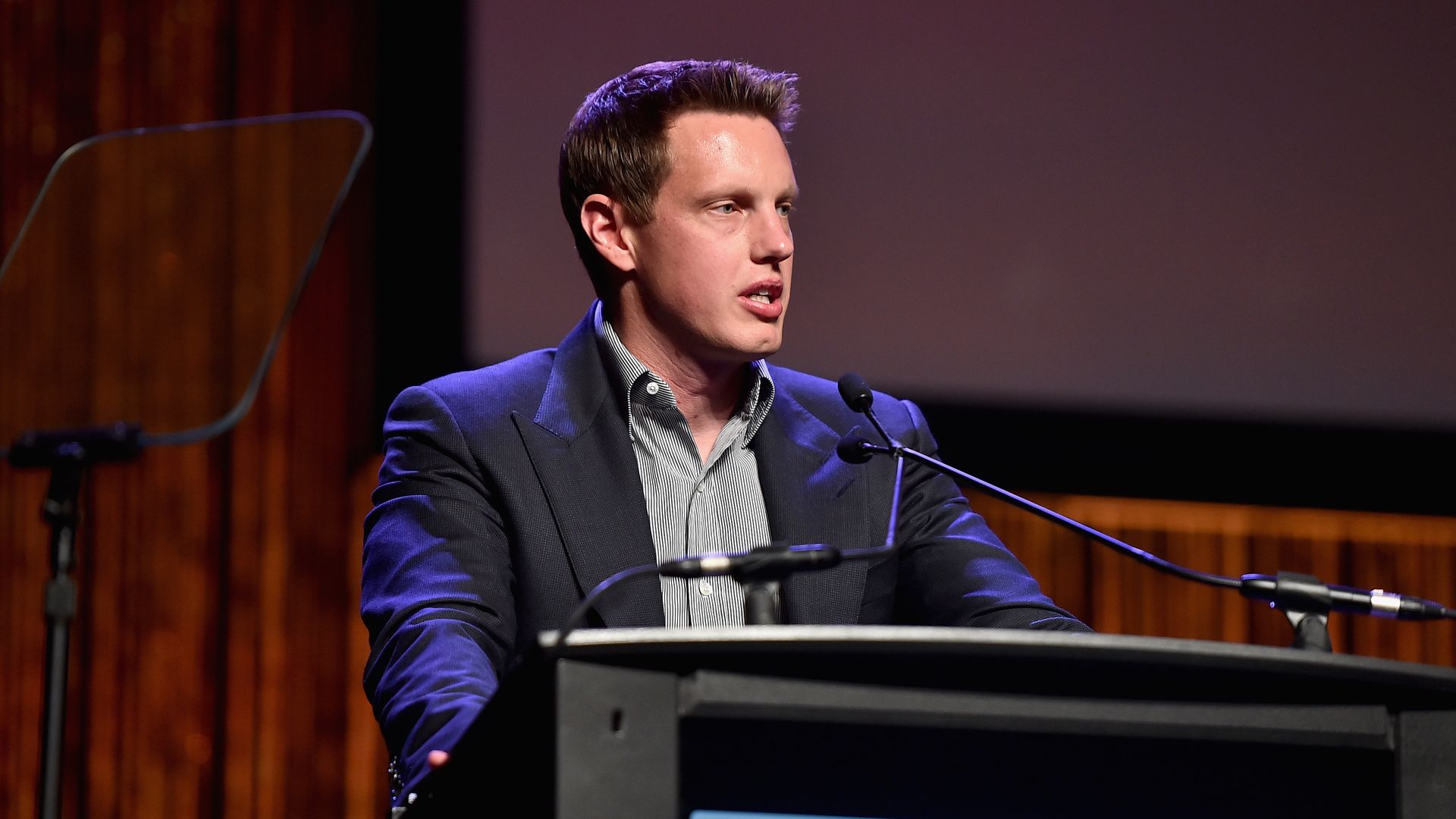 Man in dark suit and striped shirt speaking at a podium with microphones, wooden background, and large screen behind him in a formal setting.