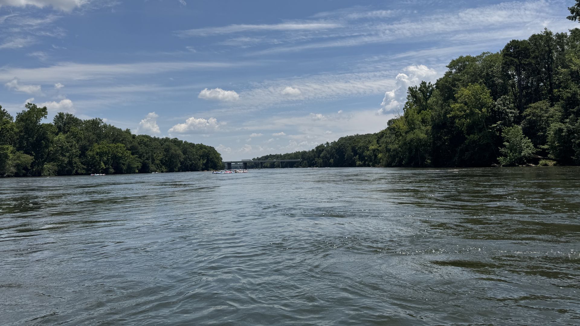 Calm river with gentle ripples, green tree-lined banks on both sides, under a blue sky with scattered clouds and a distant bridge.