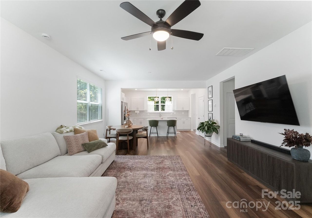 Bright open living room with white walls, a gray sectional sofa with patterned cushions, wooden floor, large rug, round dining table, green chairs, modern kitchen, and ceiling fan with light.