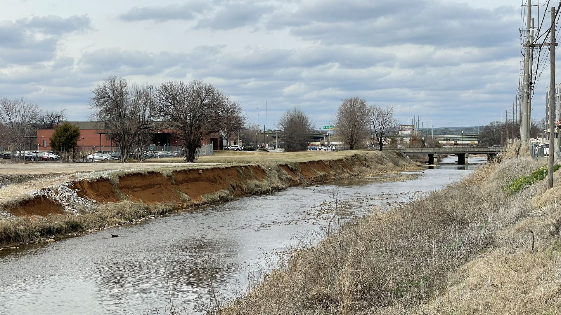 A wide river with eroded reddish banks flows through a barren, grassy area under a cloudy sky, with leafless trees, a brick building, parked cars, and a bridge in the distance.