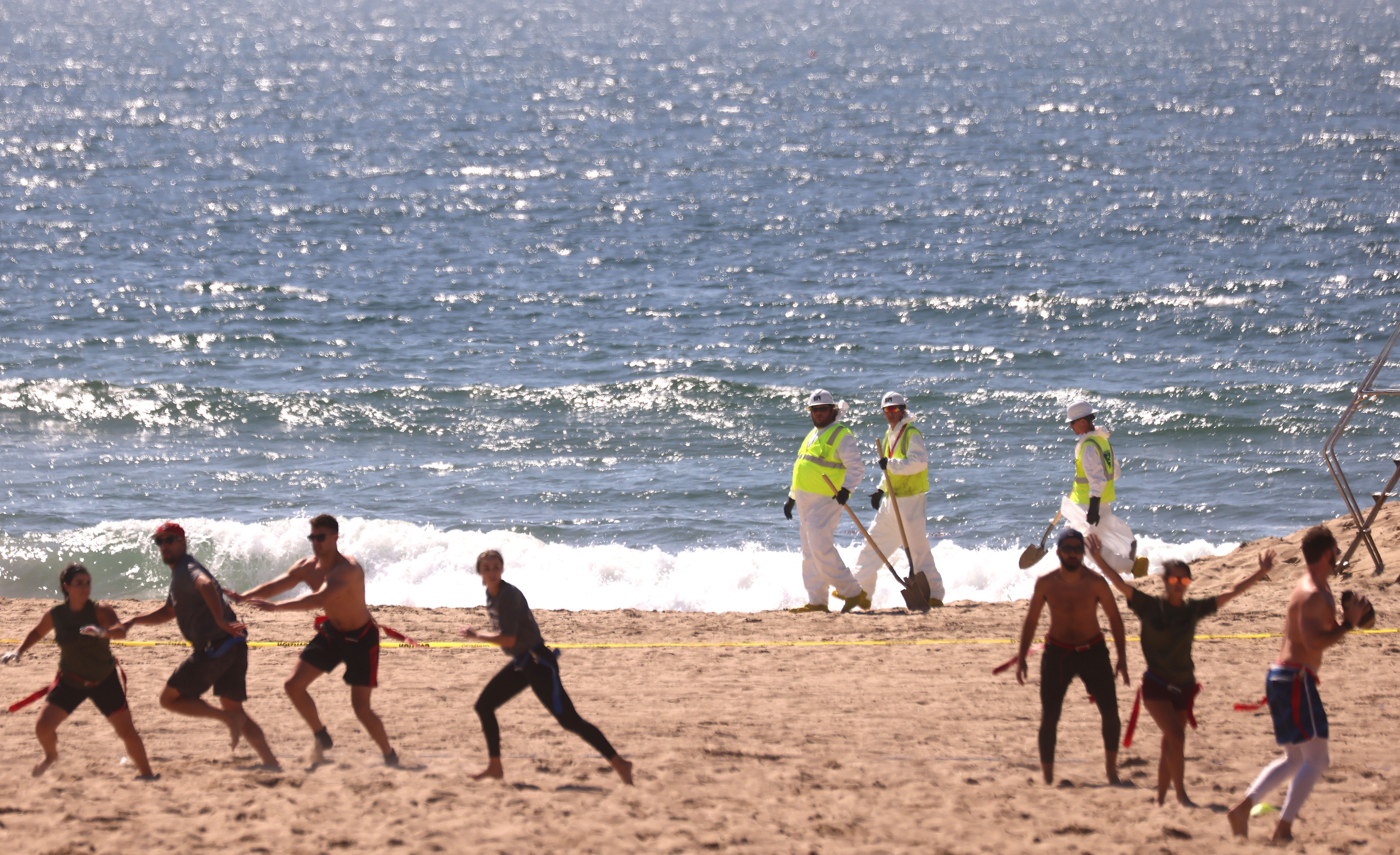  People play football as cleanup workers look for contaimination in Huntington Beach, California.  