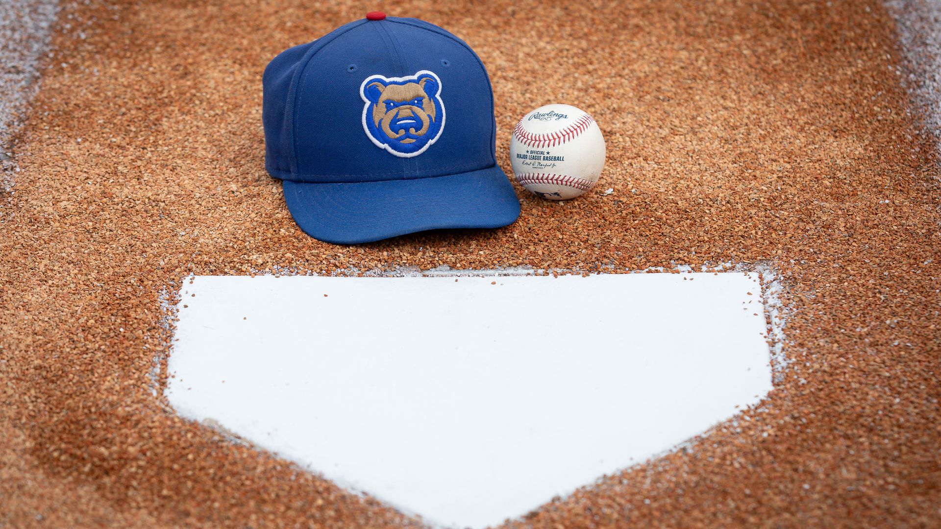 A blue baseball cap with a bear logo rests beside a baseball on brown dirt near a white home plate on a baseball field.