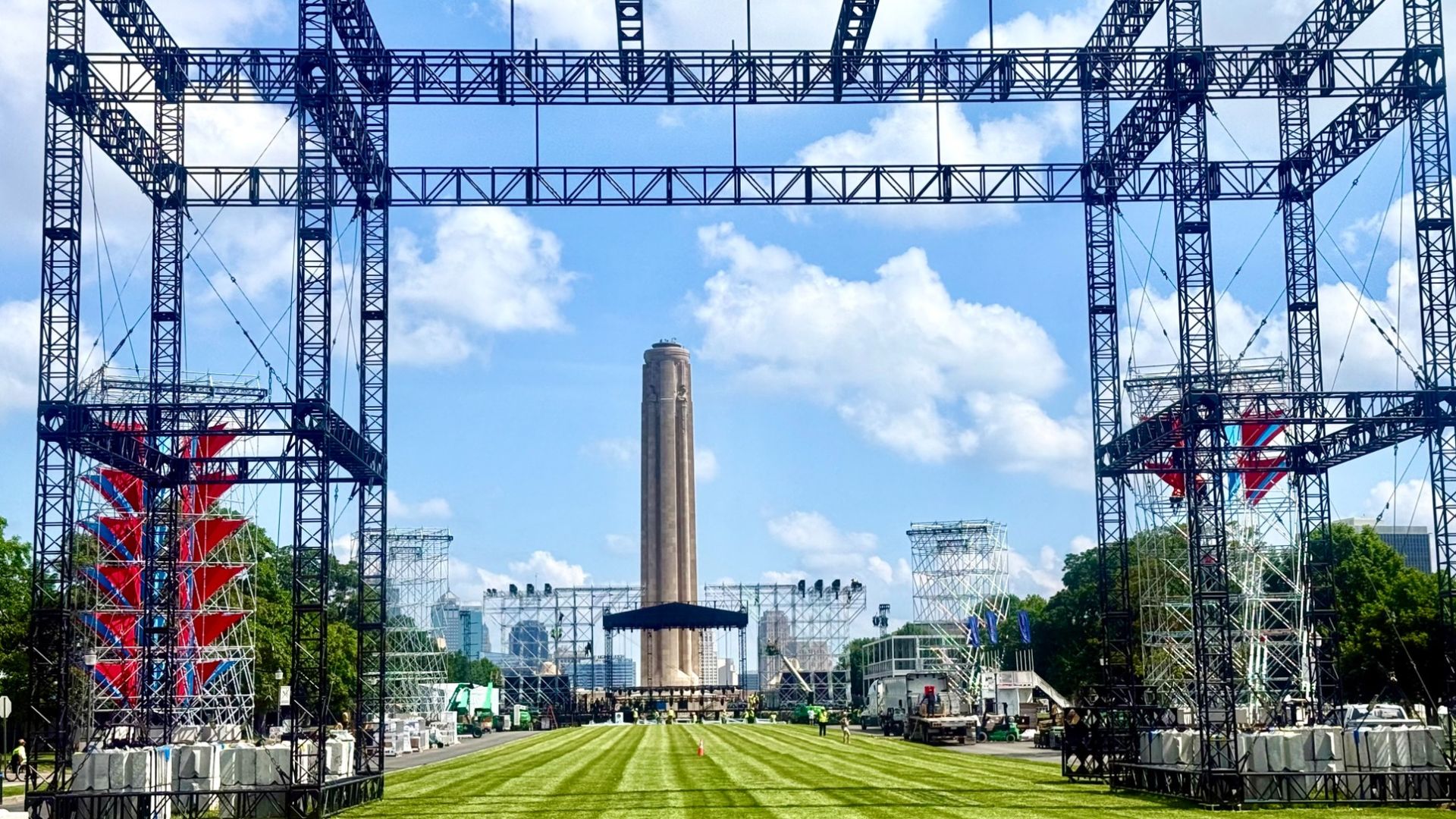 Outdoor stage framed by black metal trusses, with a tall monument at center-right. A green striped lawn leads to the stage under a blue sky with clouds; scaffolding and equipment fill the foreground.