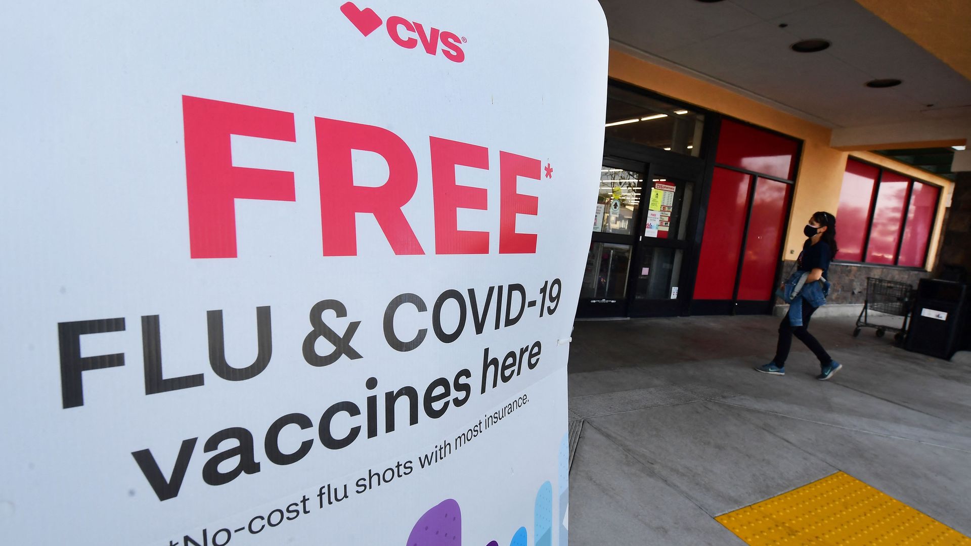 A white sign advertising free flu and COVID-19 vaccines, as a masked woman enters the CVS pharmacy.