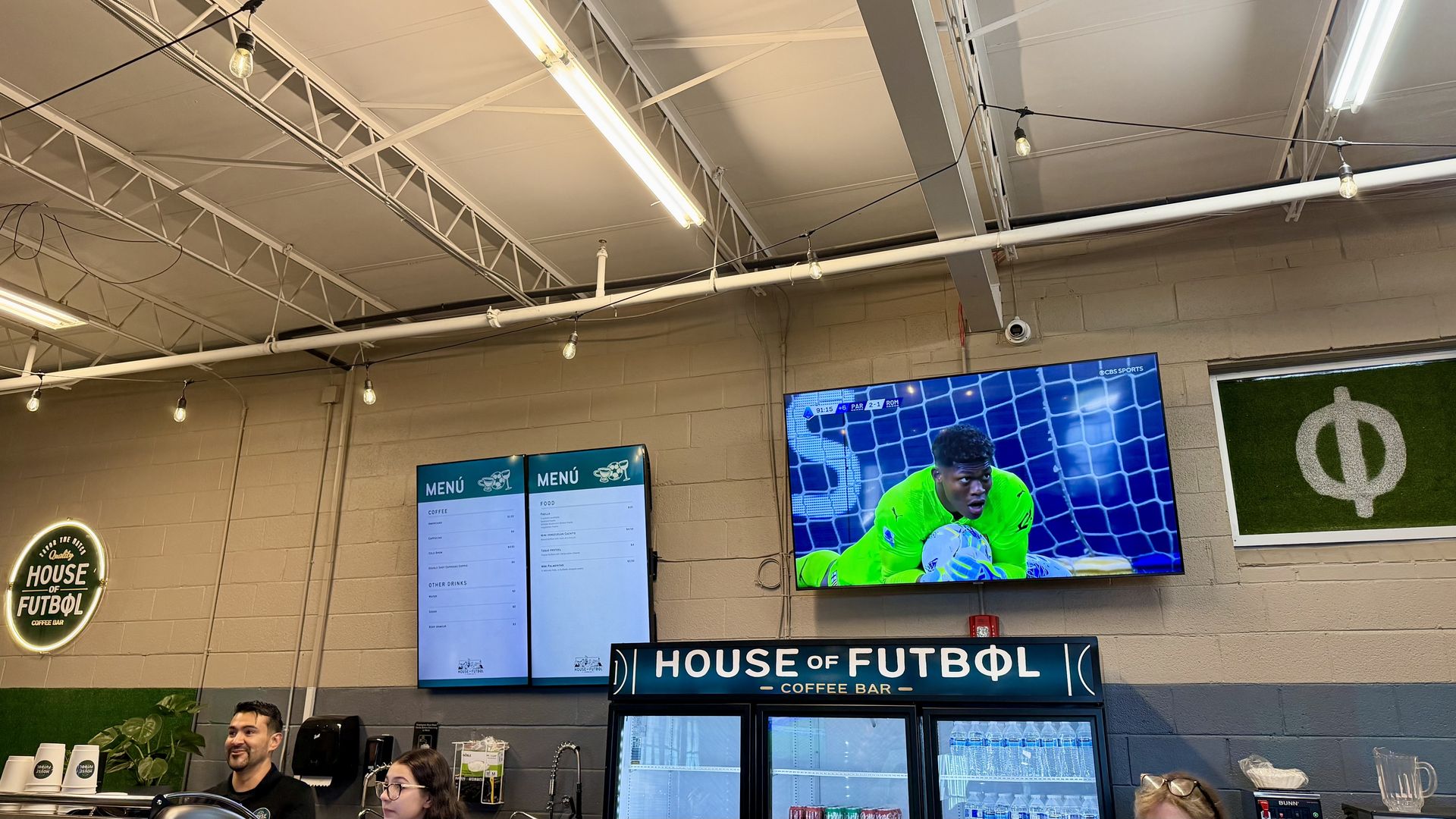 Indoor cafe with exposed white ceiling trusses and string lights. Two customers stand at the counter beneath a large TV showing a soccer match; a blue sign reads "HOUSE OF FUTBOL" and a round neon sign is on the wall.