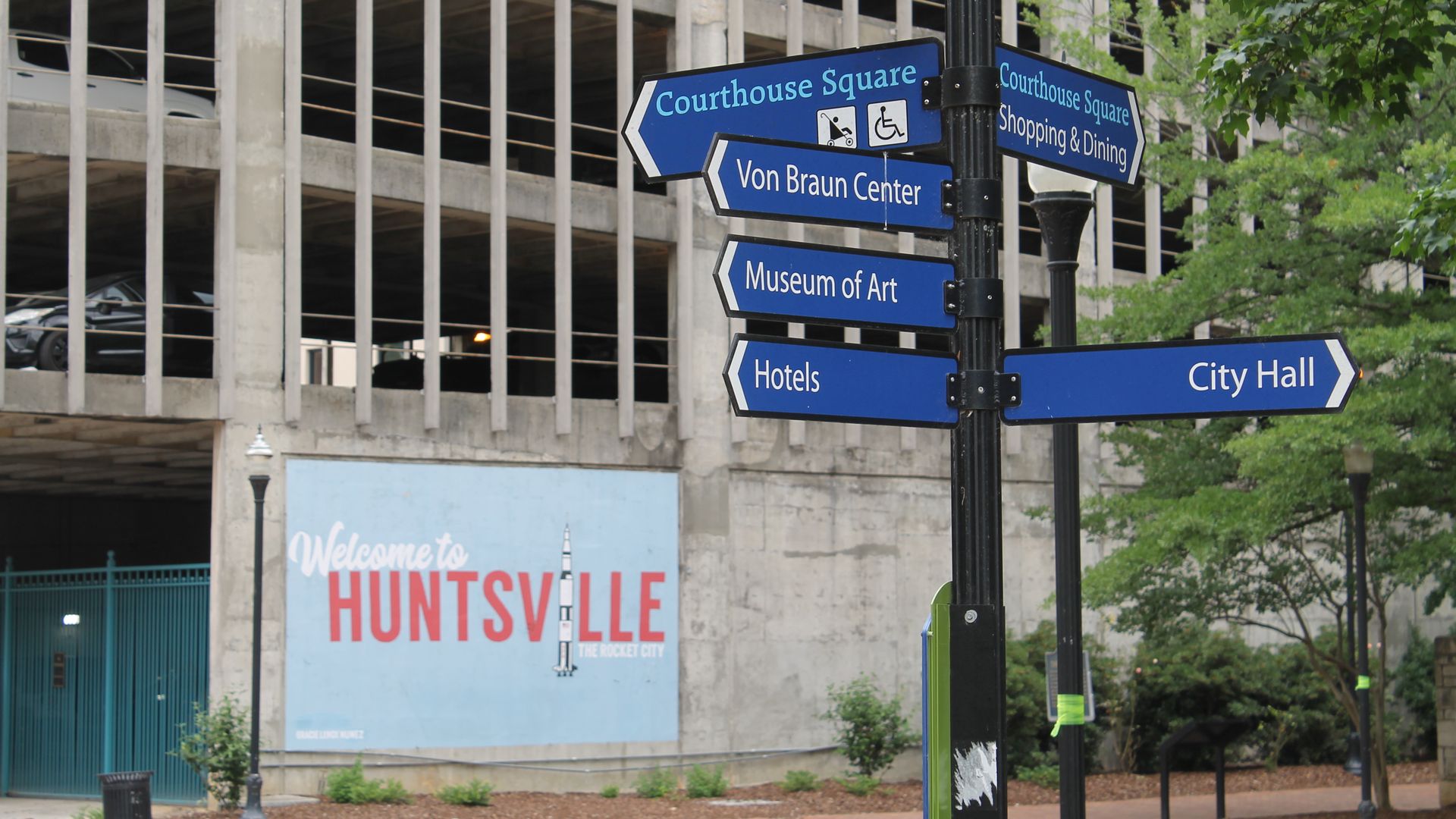 Blue street signs on a black post point to Courthouse Square, Von Braun Center, Museum of Art, Hotels, and City Hall beside a concrete parking structure; green trees frame the right, and a mural reading "Welcome to Huntsville" is below.