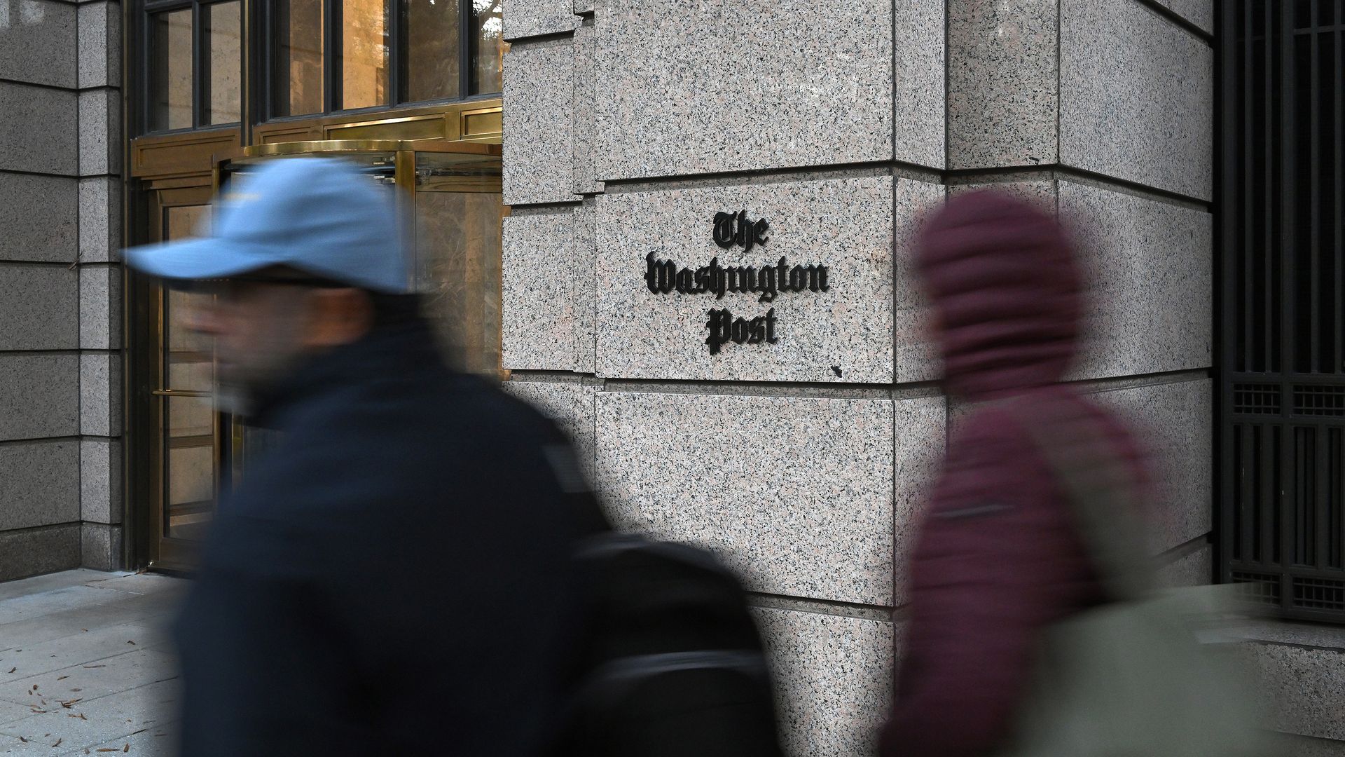 Размытые фигуры двух людей, проходящих мимо входа в здание The Washington Post с вывеской на бежевой каменной стене и вращающейся дверью на заднем плане.