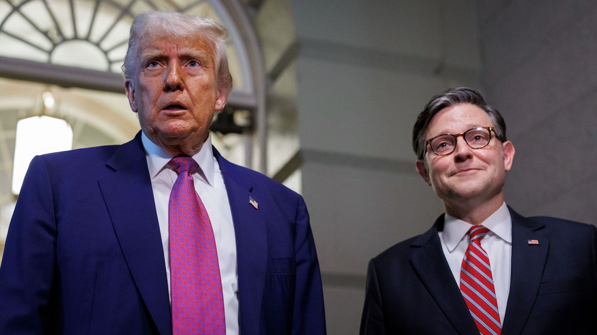 Trump and Mike Johnson standing next to each other in a white marble hallway while wearing blue suits.