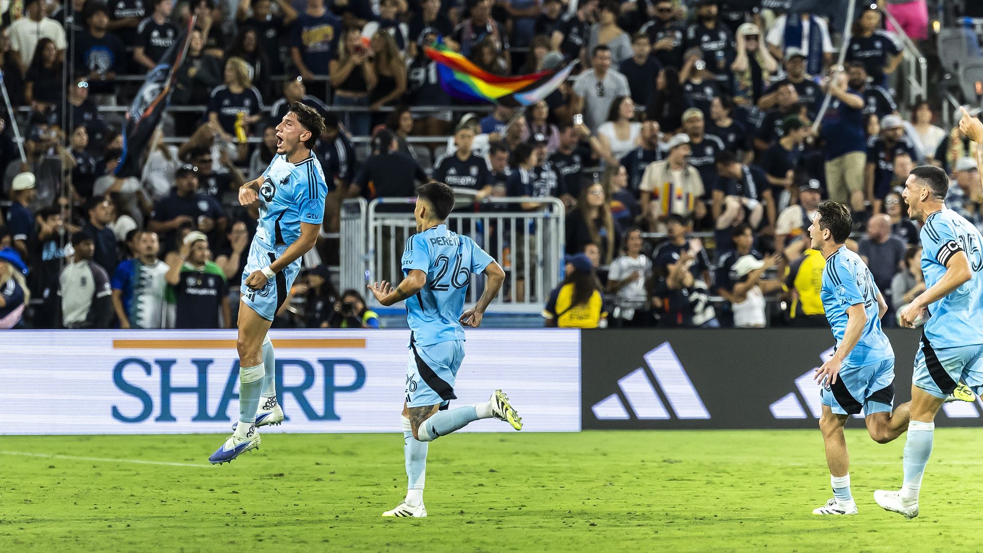 A soccer player leaps in the air in celebration after scoring a goal. Pursued by his joyful teammates in powder-blue uniforms, a quiet crowd of opposing fans watches in shock behind him.