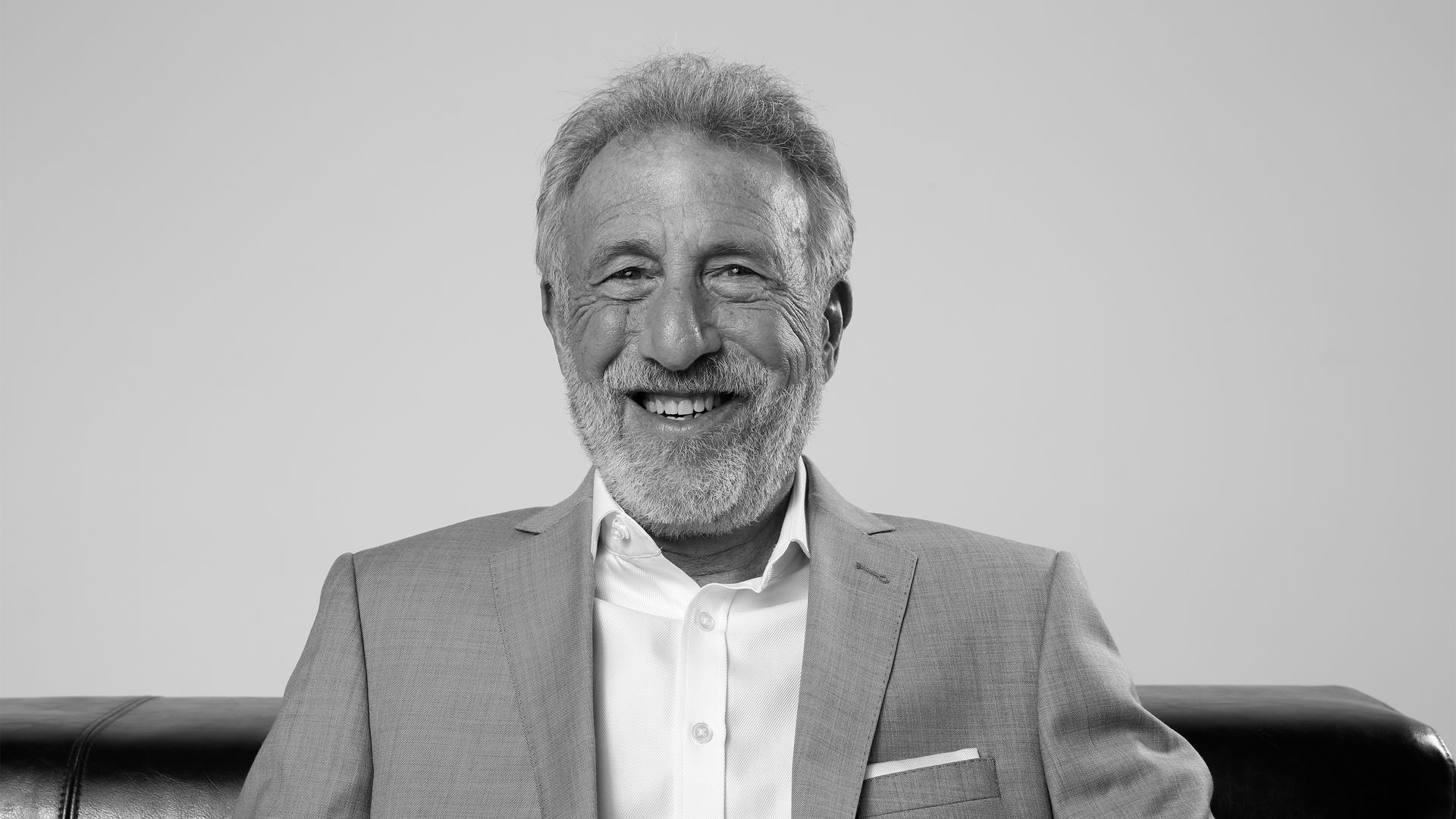 A head shot of Generation Tux founder George Zimmer, in a white button-down shirt and gray suit jacket.