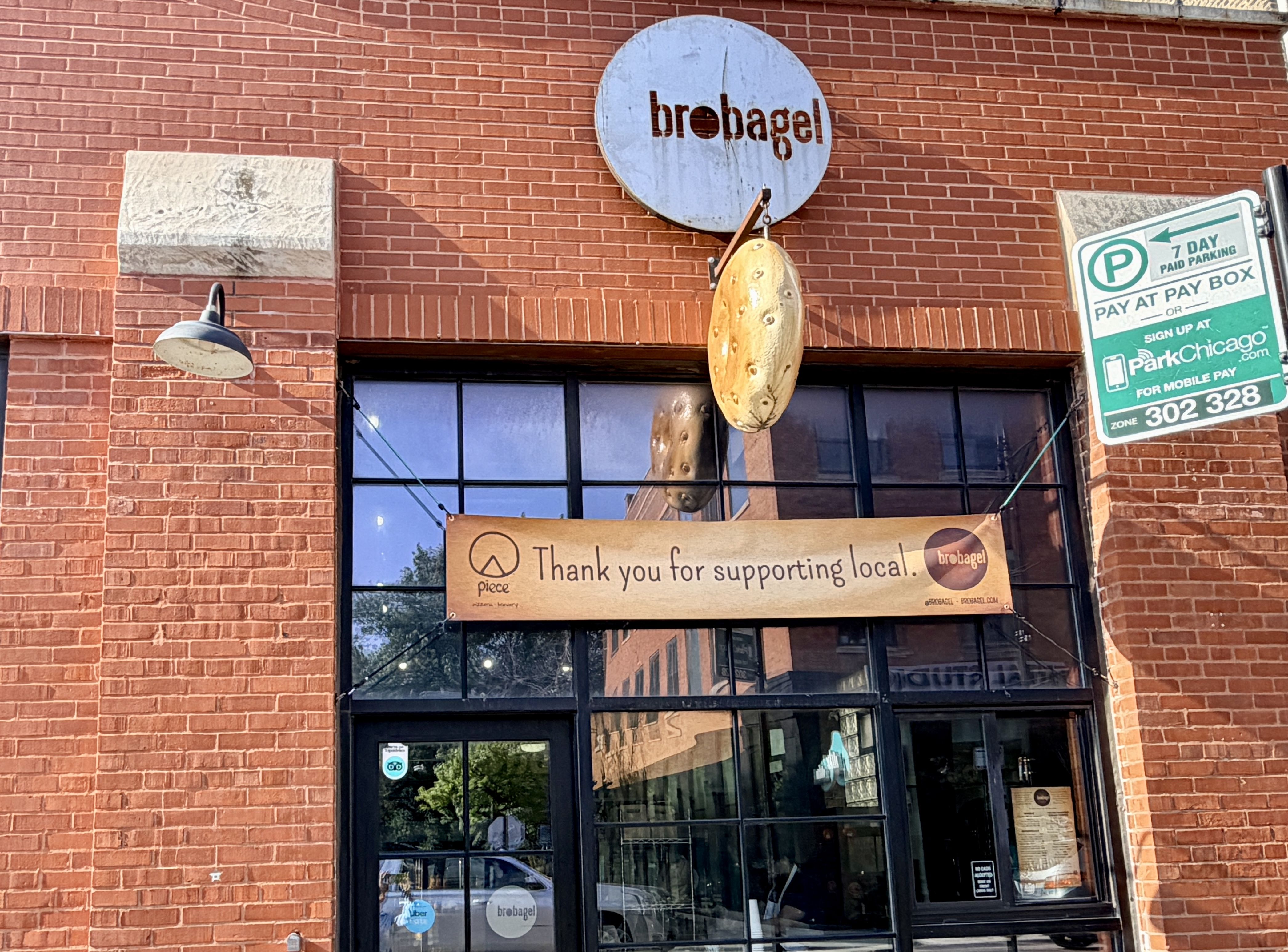 Red brick storefront with a round blue sign reading "brobagel" above a hanging bagel sculpture. To the right is a green "7 Day Paid Parking" sign; a beige banner thanks locals below.