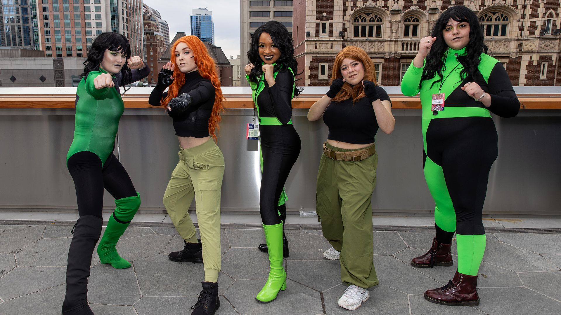 Five women stand on a rooftop in green and black cosplay gear, in a boxing pose with their hands up as if ready to fight.