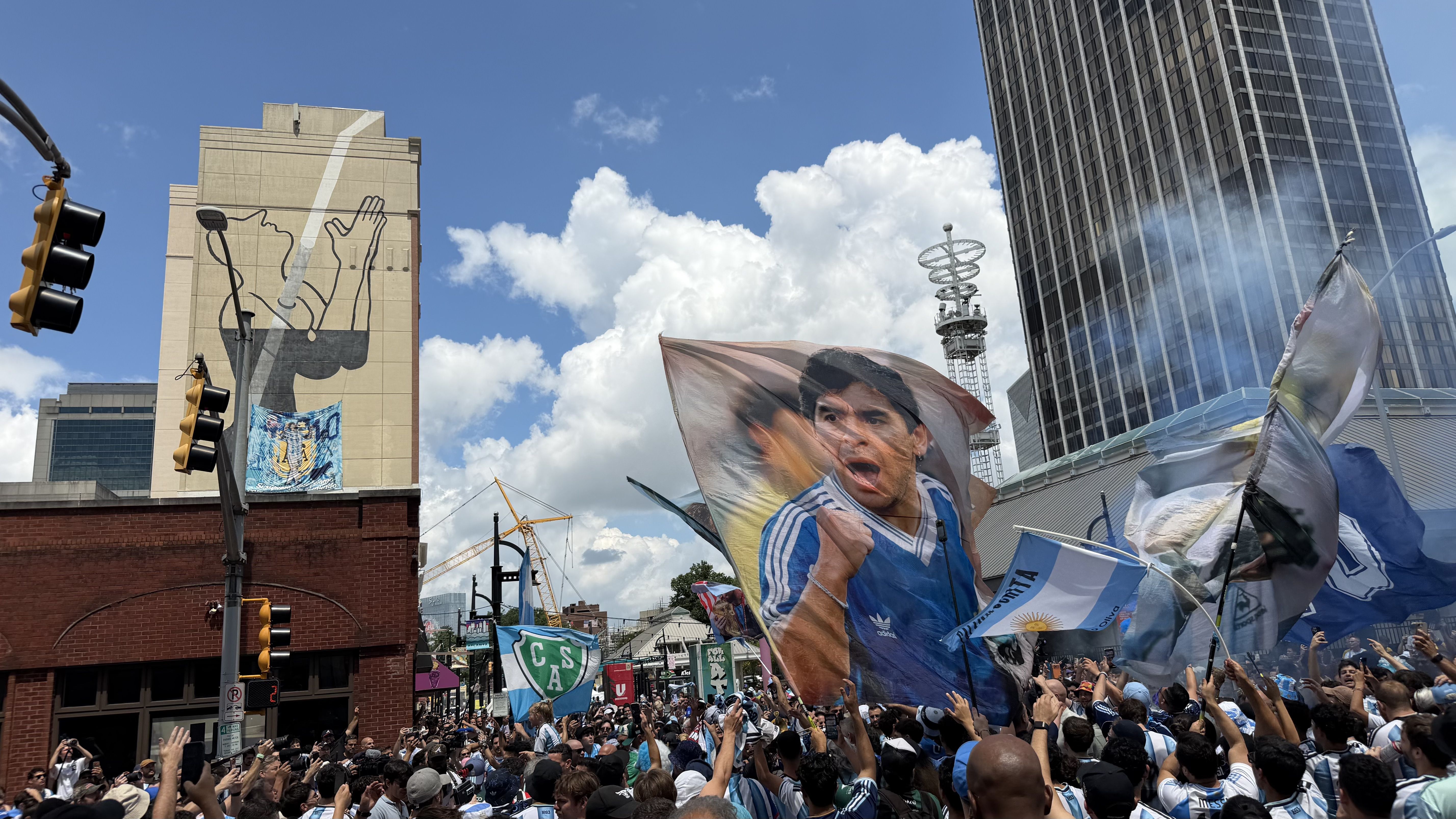 A crowded urban protest with blue and white flags and banners; a mural of a hand holding a microphone on a beige building, and tall glass skyscrapers rise under a bright blue sky.