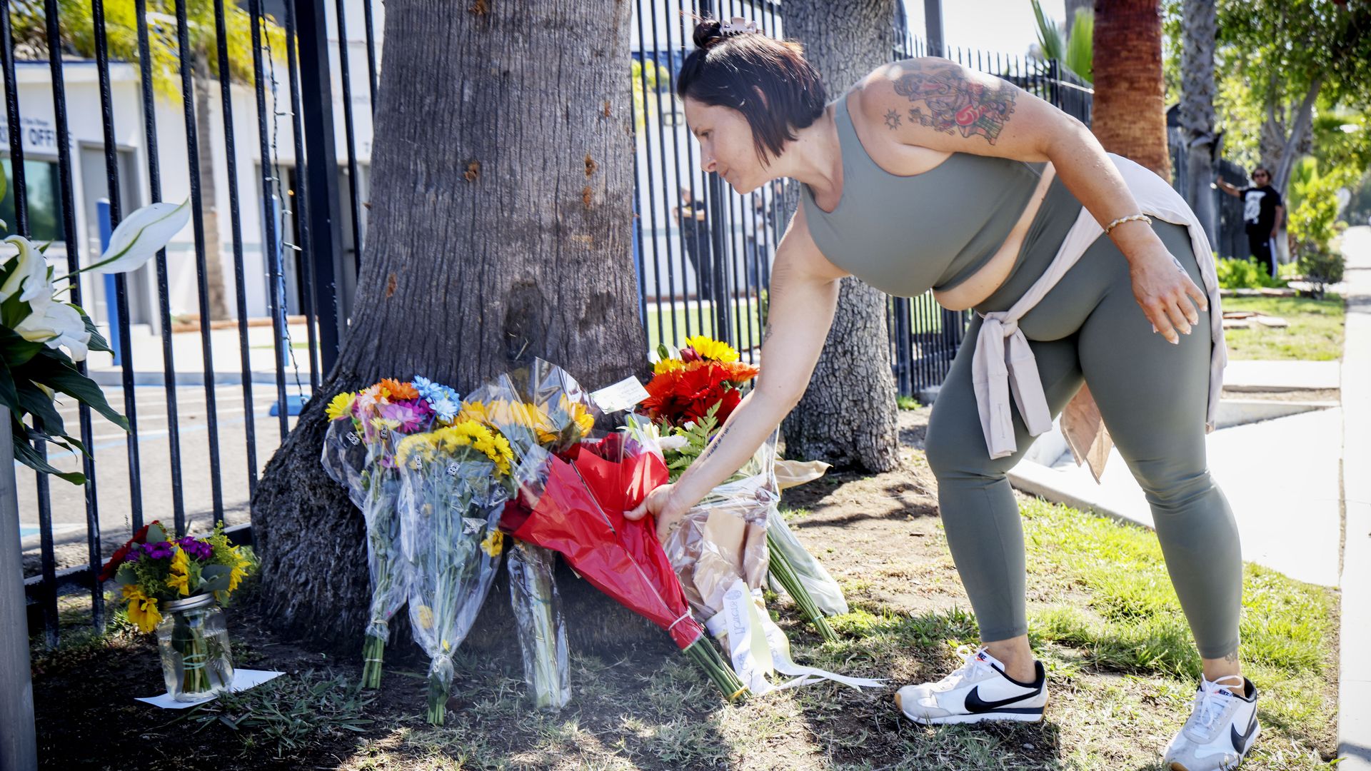 Athletic woman with a tattoo on her arm bends to arrange bright bouquets at a flower display by a black metal fence; sunny day, bystanders visible in the background.