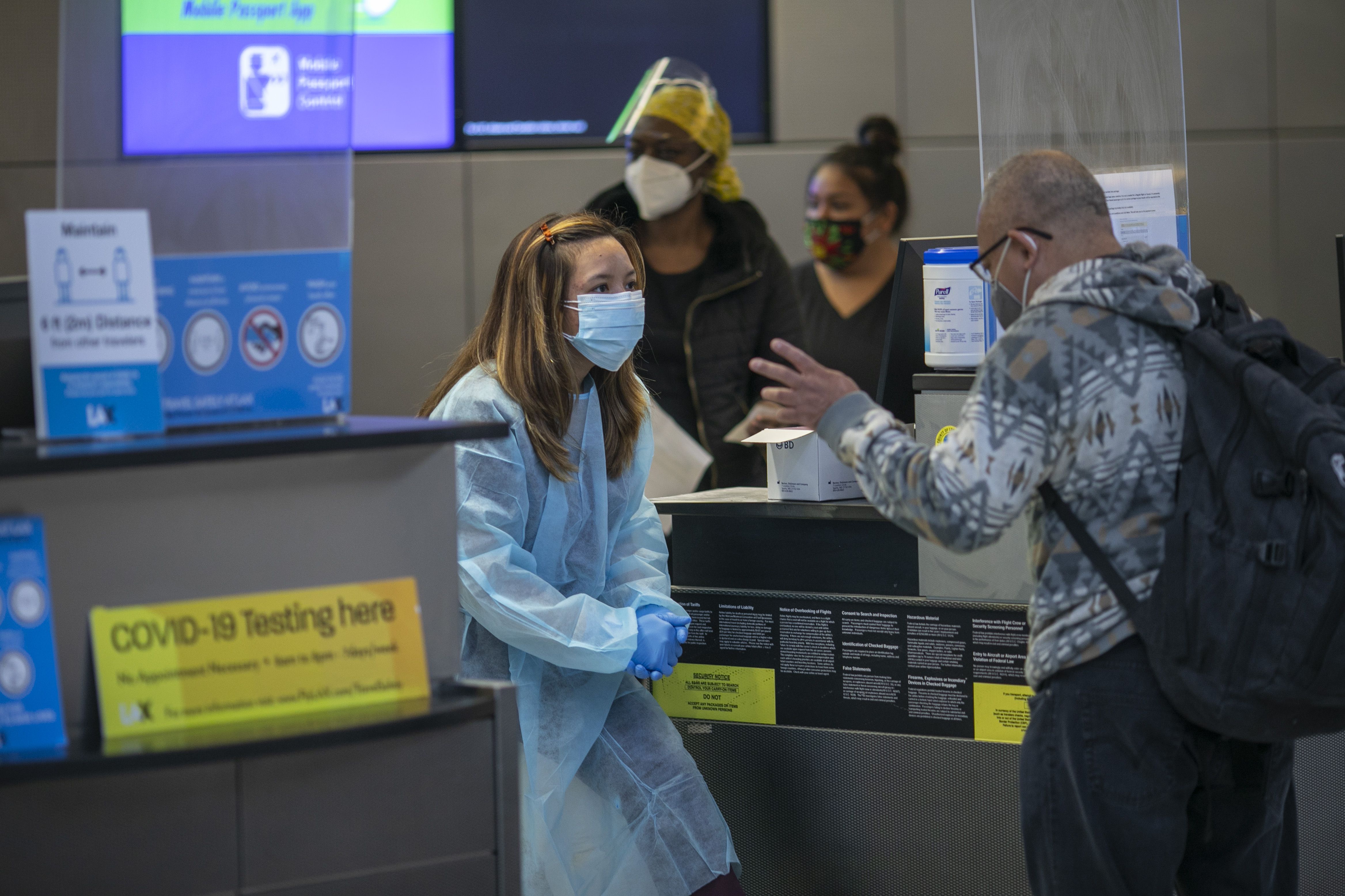 A nurse wearing a face mask and PPE stands in front of a man wearing a hoodie and backpack 