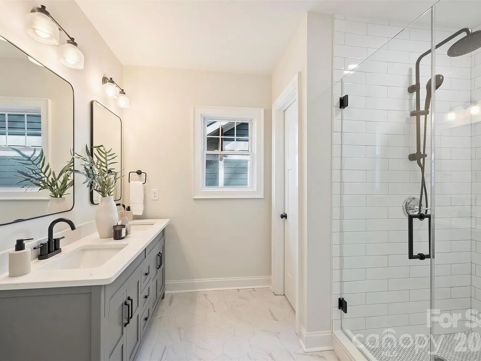 Bright modern bathroom featuring a gray double vanity, white countertop, black faucets, and two mirrors with lights. Glass-enclosed shower with white subway tile and a small plant on the counter.