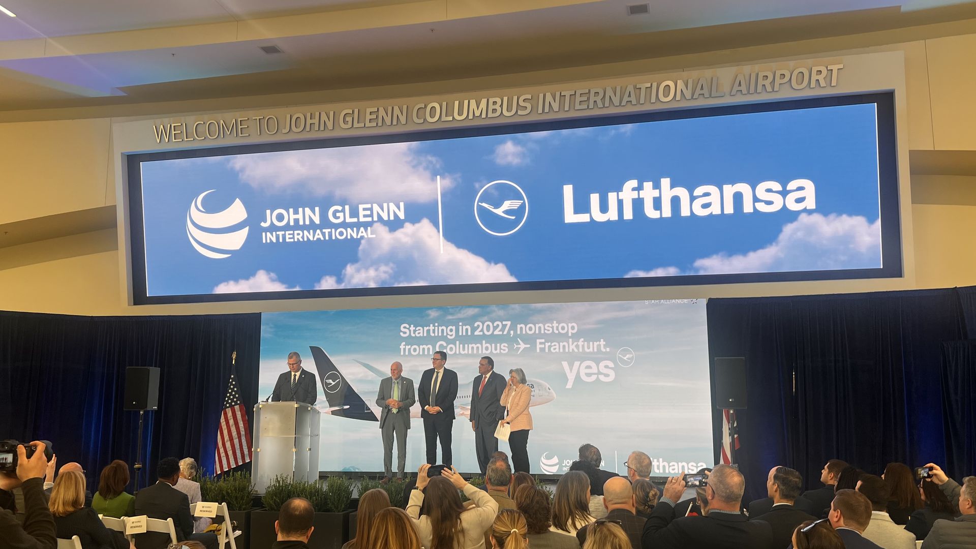 Indoor airport scene with a sign reading "Welcome to John Glenn International Airport" and logos for John Glenn International and Lufthansa; audience on chairs facing a panel.