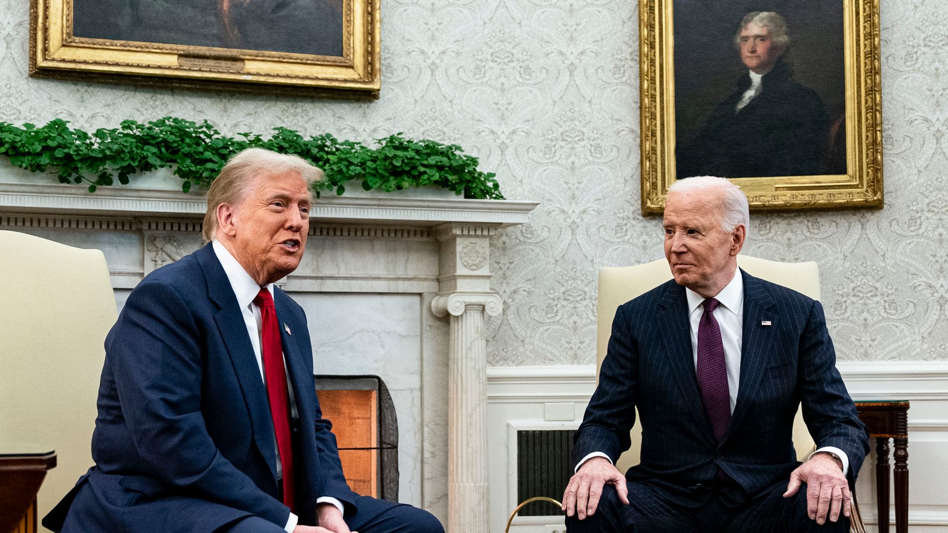 President Trump, wearing a navy suit with a US flag pin at the top of his left lapel, white shirt and red tie, smiles as he speaks in the White House Oval Office as President Biden, wearing a black suit, white shirt and purple tie looks on.