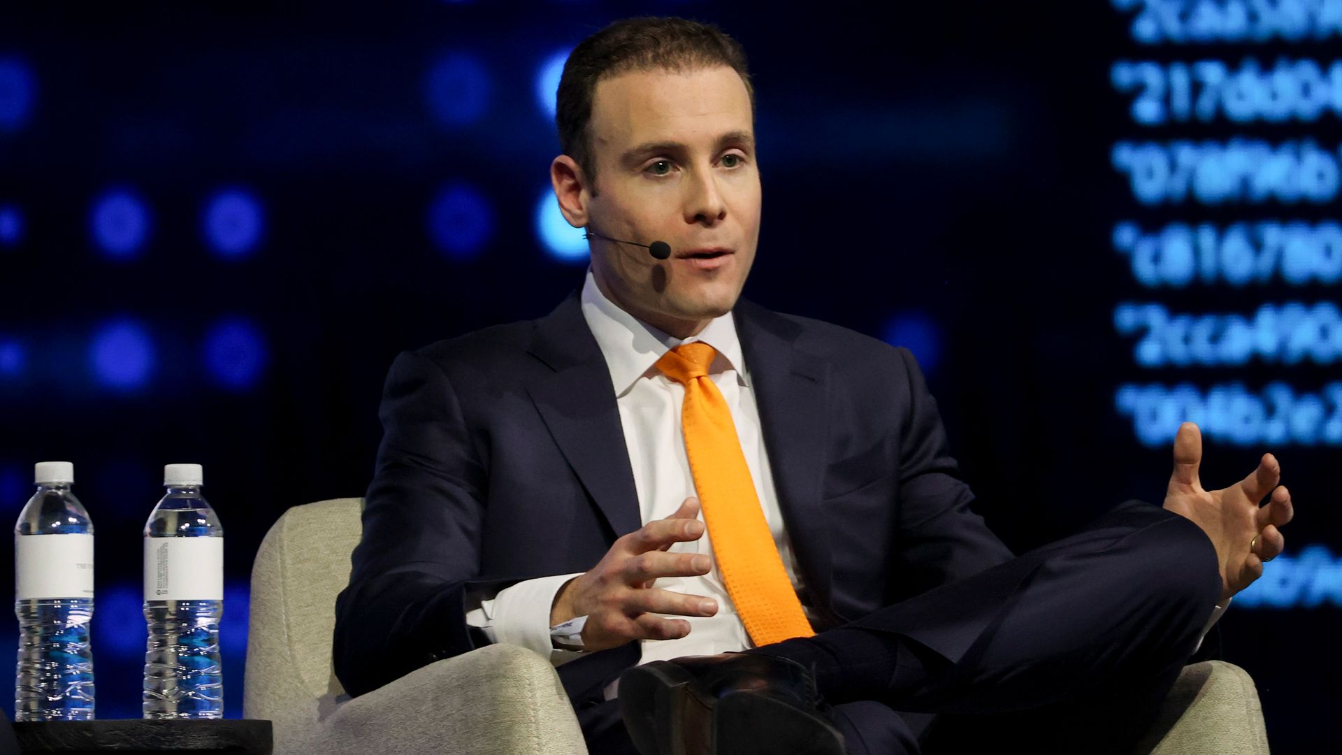 A person in a dark suit and orange tie speaks on a stage, wearing a headset microphone and gesturing with his hands. Water bottles sit on a table nearby, blue stage lights in the background.