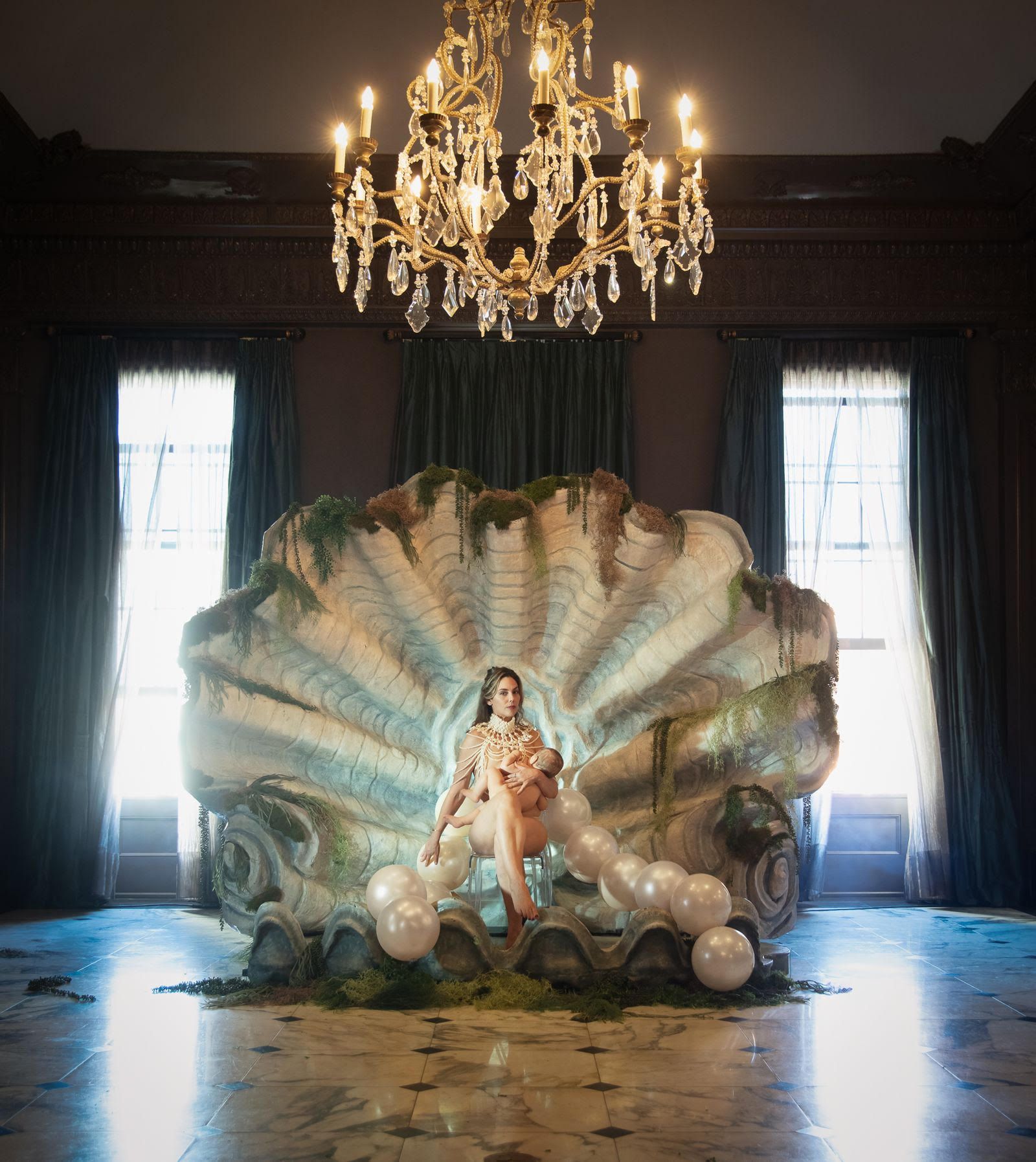 Woman breastfeeding a baby while sitting on a chair inside a giant, decorated clam shell with greenery and white balloons, beneath an ornate crystal chandelier in a dim room.