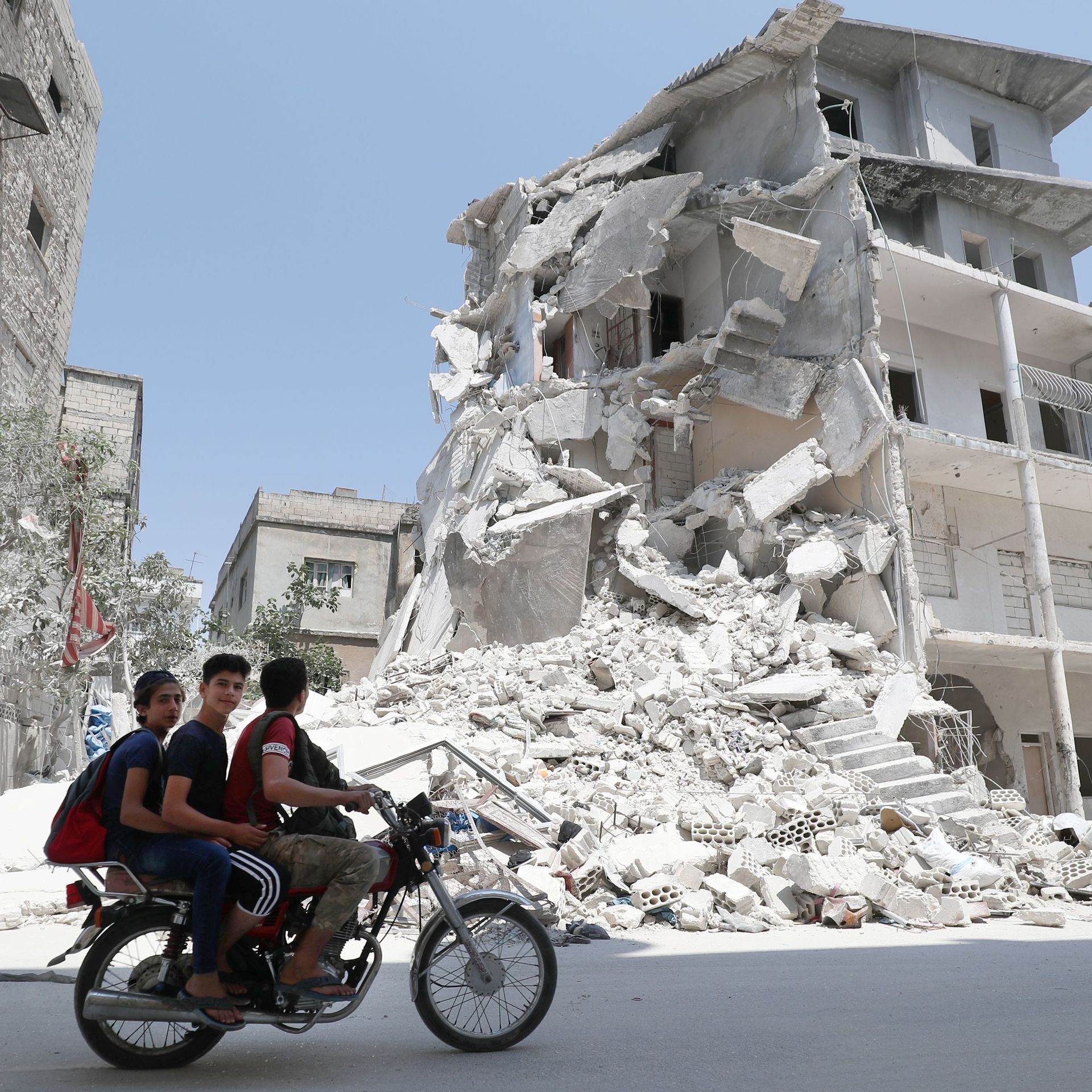 Men on a motorcycle ride by an airstrike-damaged building